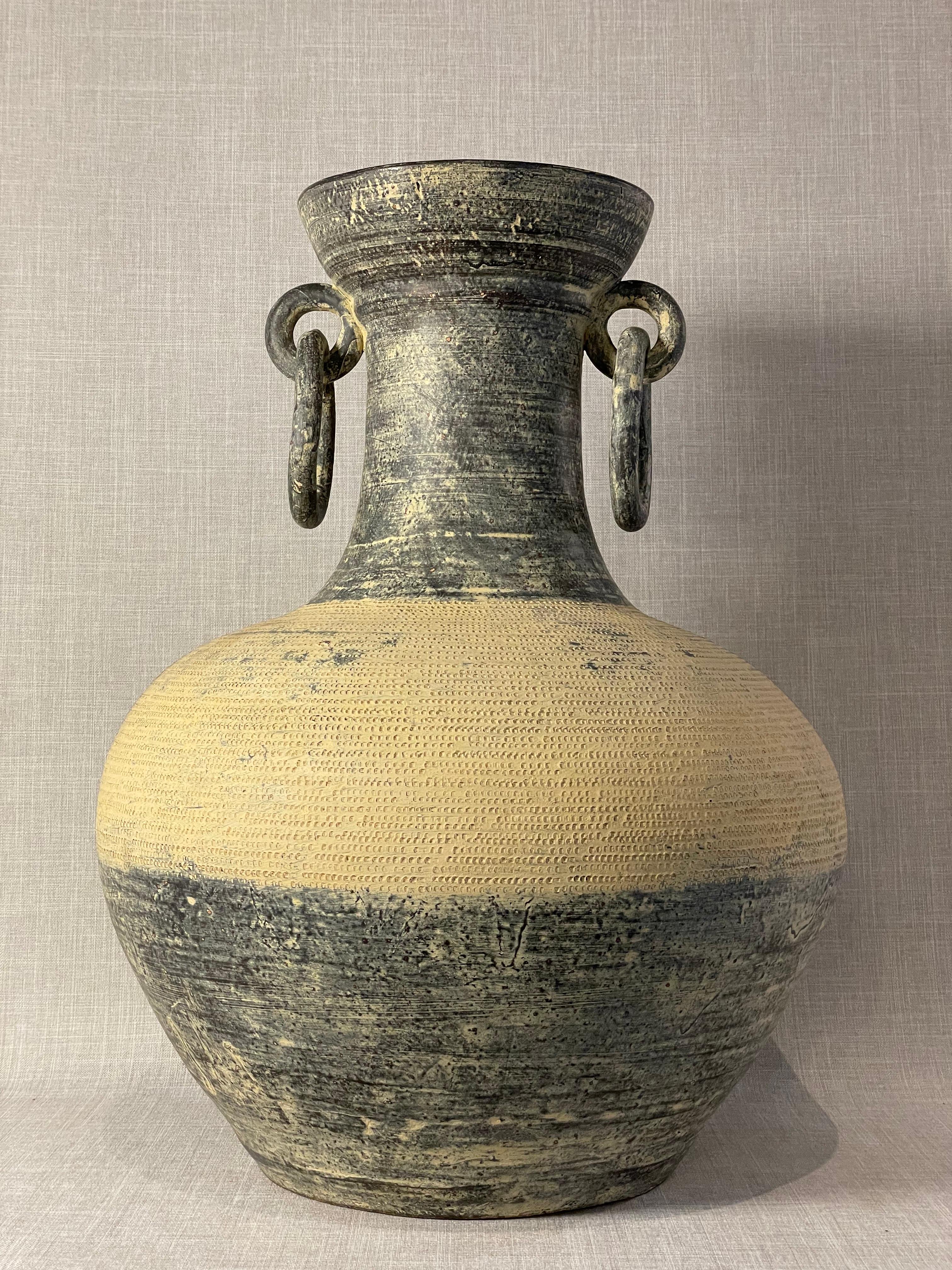Questo vaso monumentale in Studio Pottery è un pezzo scultoreo che unisce la forma antica all'artigianato moderno. La sua presenza imponente è rafforzata da un corpo robusto e arrotondato che si assottiglia con grazia in un collo alto e svasato,