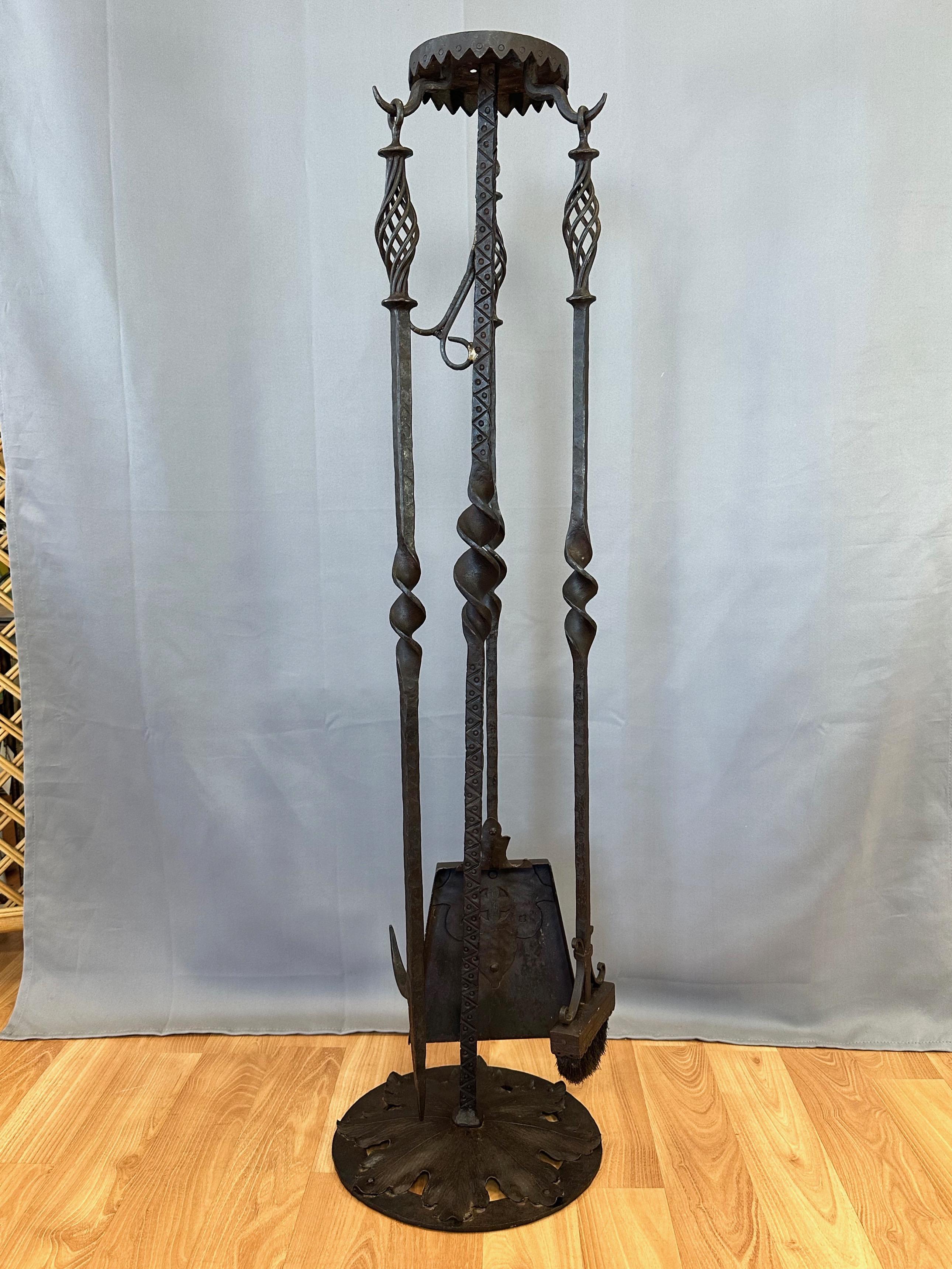 An uncommonly large and likely bespoke circa 1900 Tudor Arts and Crafts period four-piece wrought iron fireplace tool set.

Monumental hand-wrought and hand-worked set features three tools that hang from a nearly four-foot-tall stand, with a very