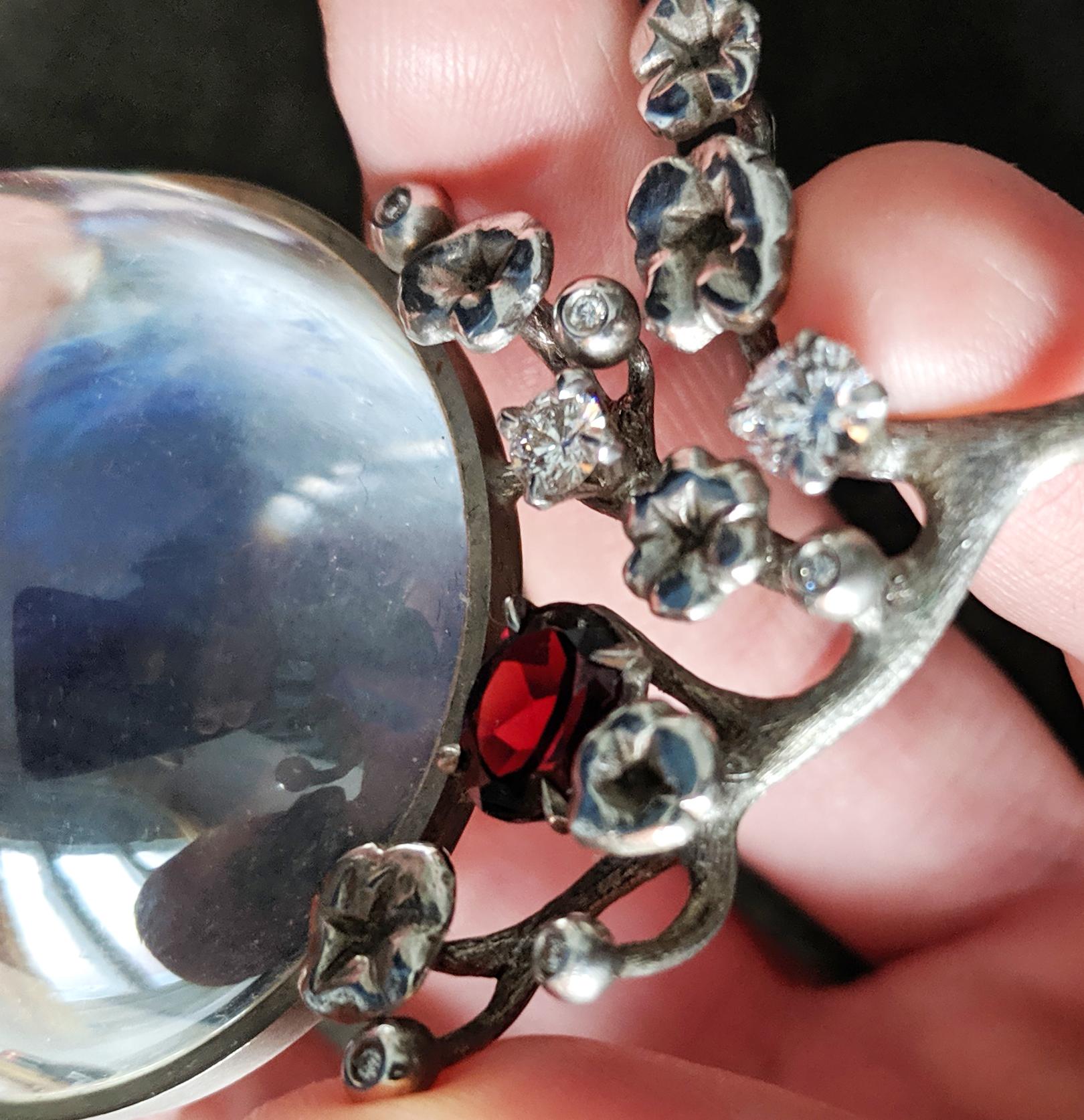 Moon Pendant is a stunning piece of jewelry crafted by an artist using 14 karat white gold and sterling silver with a large rock crystal lens, diamonds (1.5 carats), and garnet (6x8 mm). It is a limited edition work that is handcrafted.

This