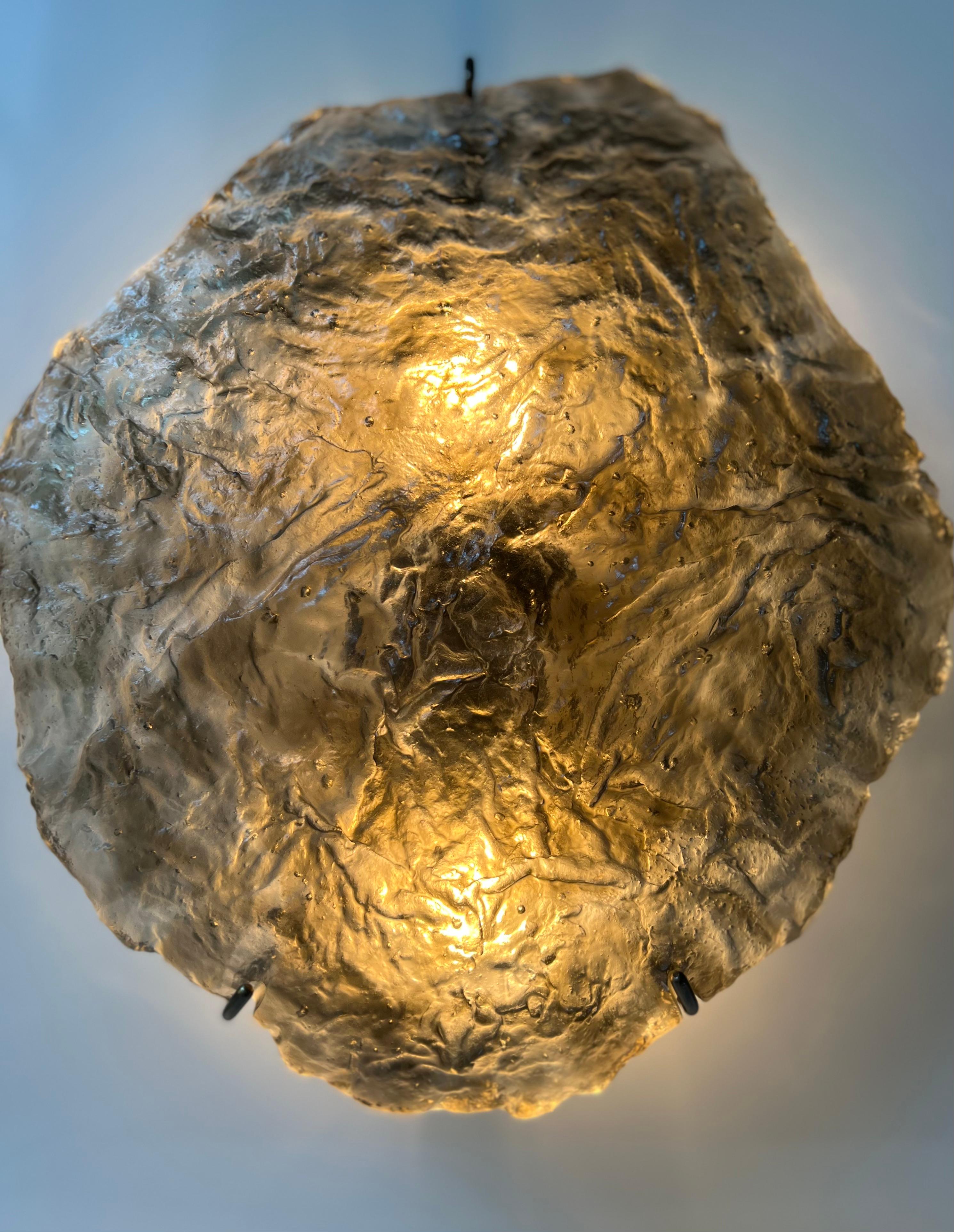 This hand-sculpted glass sconce has two regular bulbs. The installation is simple where a metal plate attaches to a wall. Three metal hooks hold the glass itself. The glass is cast in olive color in a unique technique practiced by the artist,