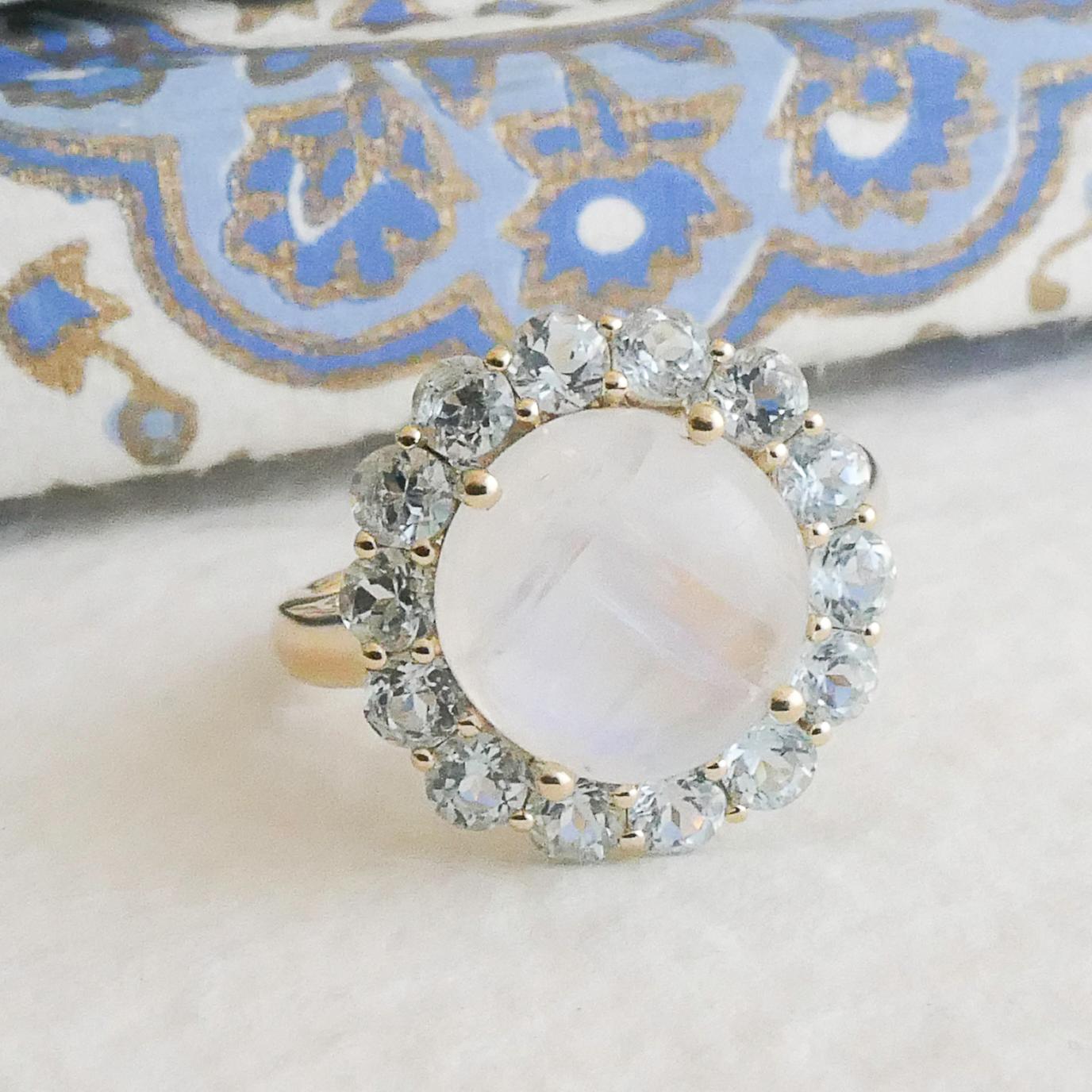 Cette bague enchanteresse présente en son centre une pierre de lune lumineuse de 11 mm, qui irradie un jeu captivant de lumière et de teintes douces. La pierre de lune est entourée d'un halo de topazes bleues étincelantes de 3 mm, méticuleusement