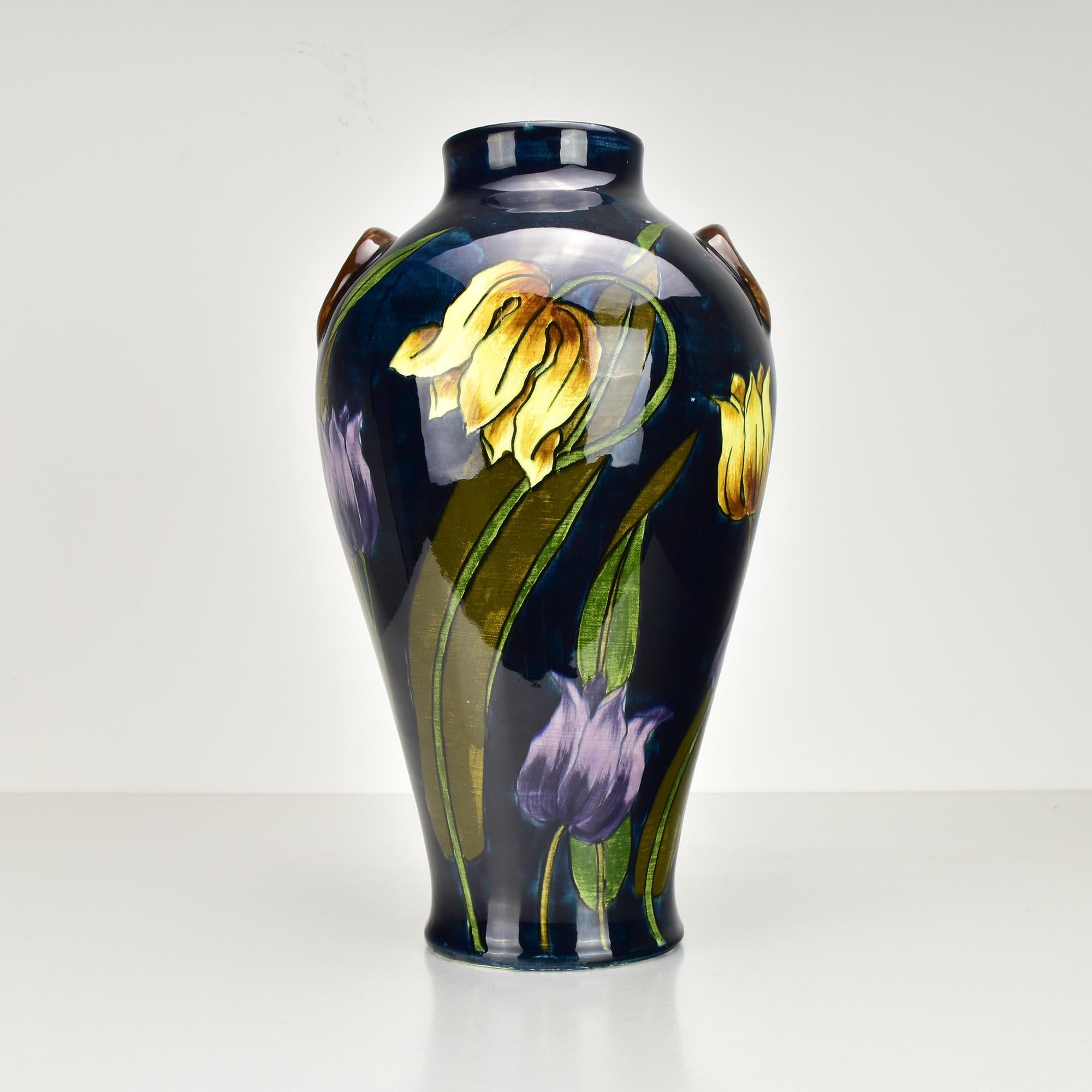 This stunning Art Nouveau ceramic vase, attributed to Moorcroft, features an exquisite hand-painted tulip decor. The deep, glossy glaze accentuates the vibrant yellows and purples of the floral design, creating a captivating contrast on the dark