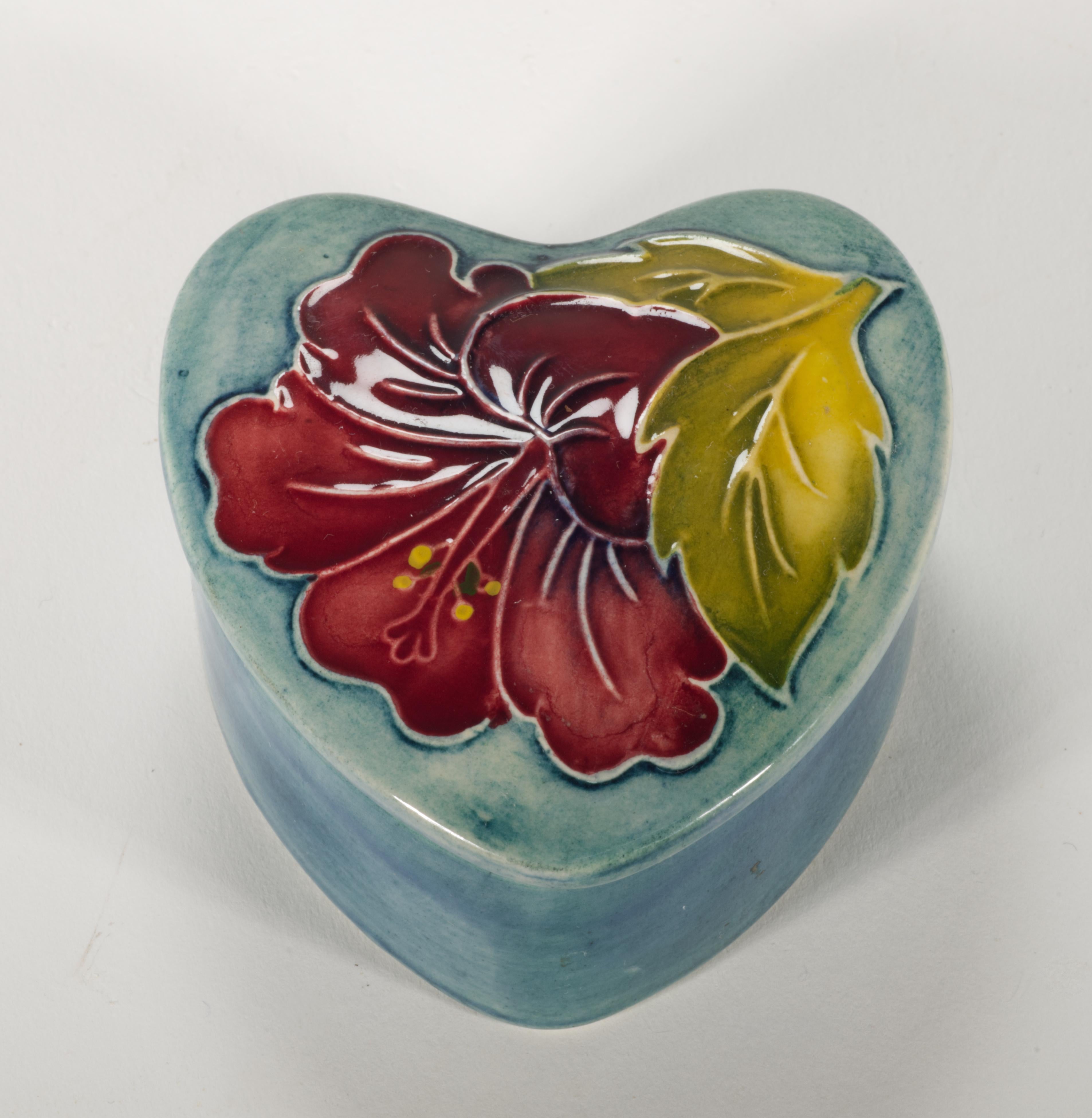 Die bezaubernde Vintage-Keramikdose mit Deckel zeigt das Dekor Hibiskus mit einer auffälligen roten Blüte und gelb-grünen Blättern, die von Hand auf ein leicht erhöhtes Relief gemalt wurden. Die weiche Form der Blume ergänzt die herzförmigen Linien