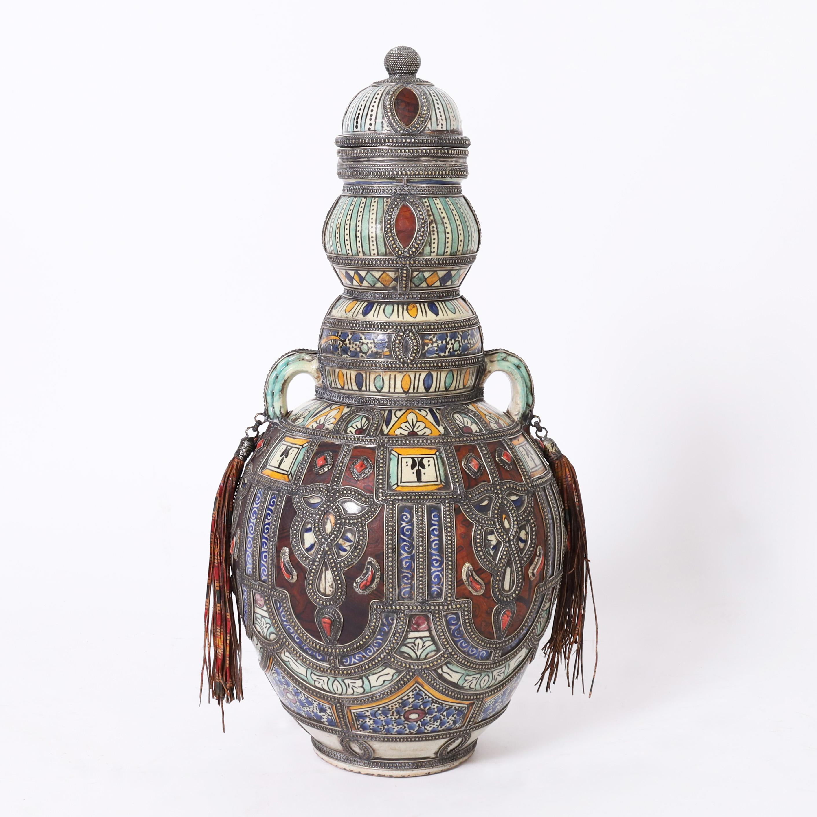 Impressive vintage pair of Moroccan lidded urns handcrafted in terra cotta decorated and glazed in distinctive mediterranean color, featuring jewelry like handcrafted metalwork with semiprecious stones and leather panels and tassels. 