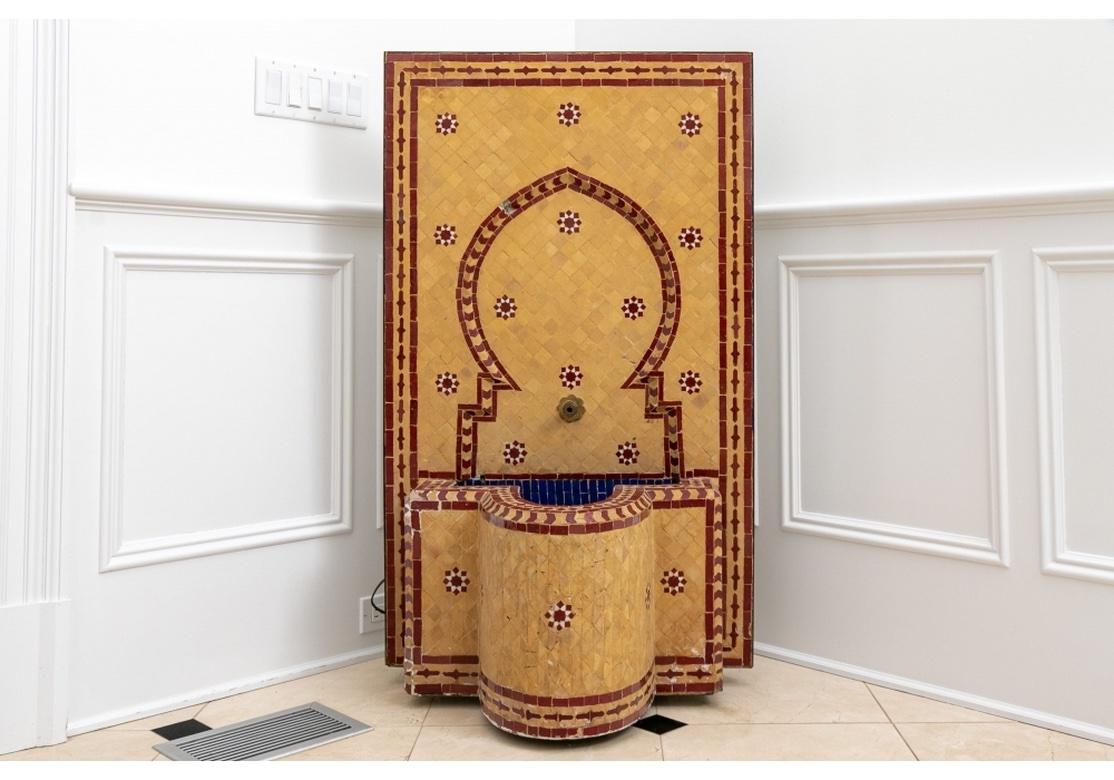 A colorful and artistic fountain in deep red and pale tan ceramic tiles with an ogival arch and rosette motifs on the back and a projecting blue tiled well below, the whole affixed to a cast stone or cement backing. With a brass spout and electric