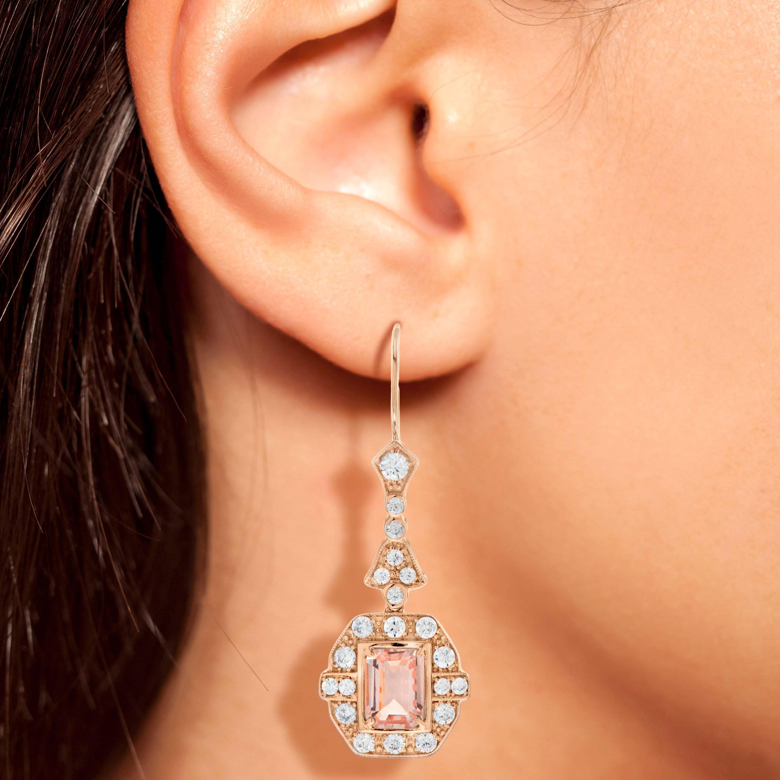 Aggiungi un tocco di glamour vintage alla tua collezione con questi orecchini pendenti mozzafiato in Morganite e diamanti in stile Art Déco. Le morbide sfumature rosa pesca delle pietre centrali di Morganite taglio smeraldo sono completate da