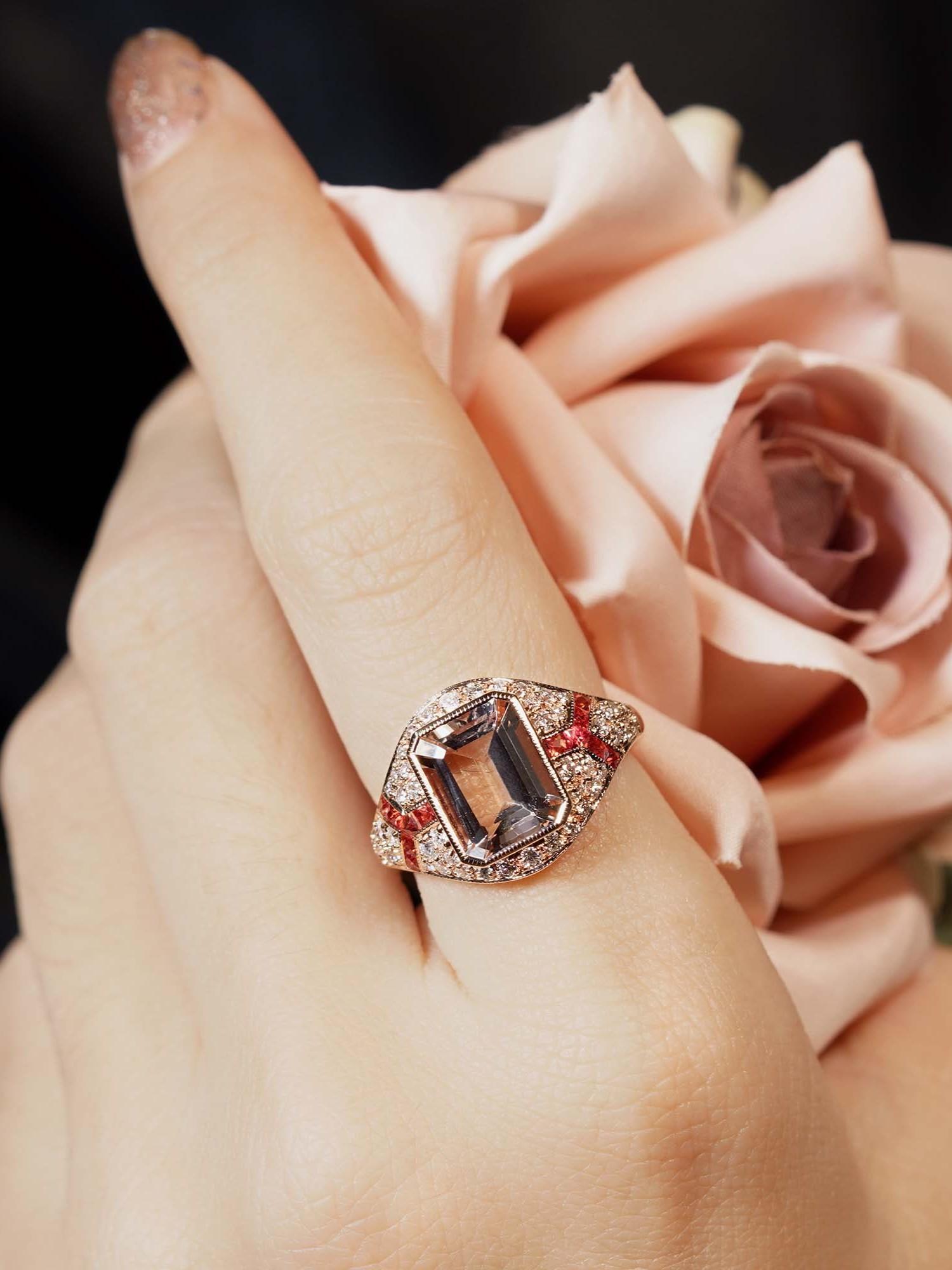 Uno splendido centro di morganite taglio smeraldo che brilla di sfumature morbide e romantiche. Affiancato da disposizioni lineari di zaffiri arancioni sia a sinistra che a destra, il design aggiunge un vibrante contrasto. Le spalle dell'anello sono