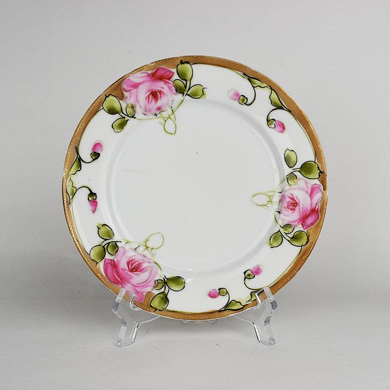 A small round ceramic plate with a pink and green floral design around the edges. The center of the plate is a crisp white glazed ceramic with slight cracquelure. The edges feature hand painted pink roses with green foliage. A raised pattern of dots