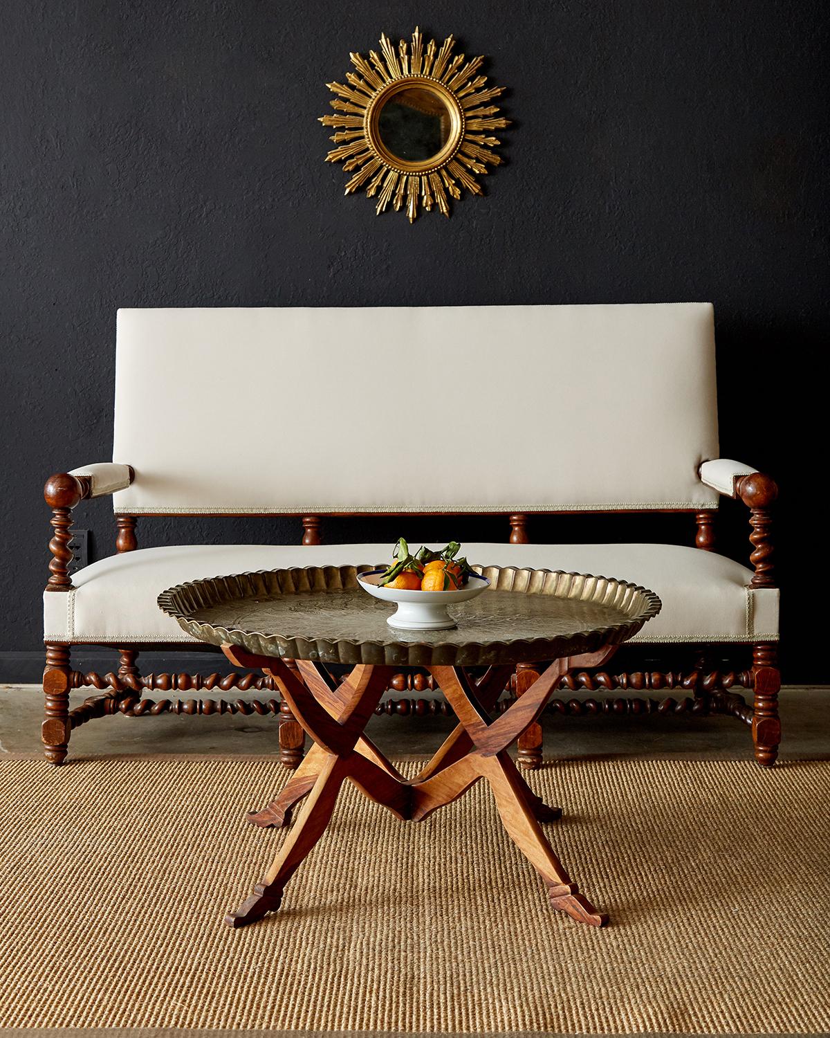 Chic Moroccan brass tray table featuring a folding mahogany base with figural arms and legs holding the large brass tray top. The Moorish Moroccan brass tray has a floral motif sunburst design on top and a traditional scalloped edge. The custom