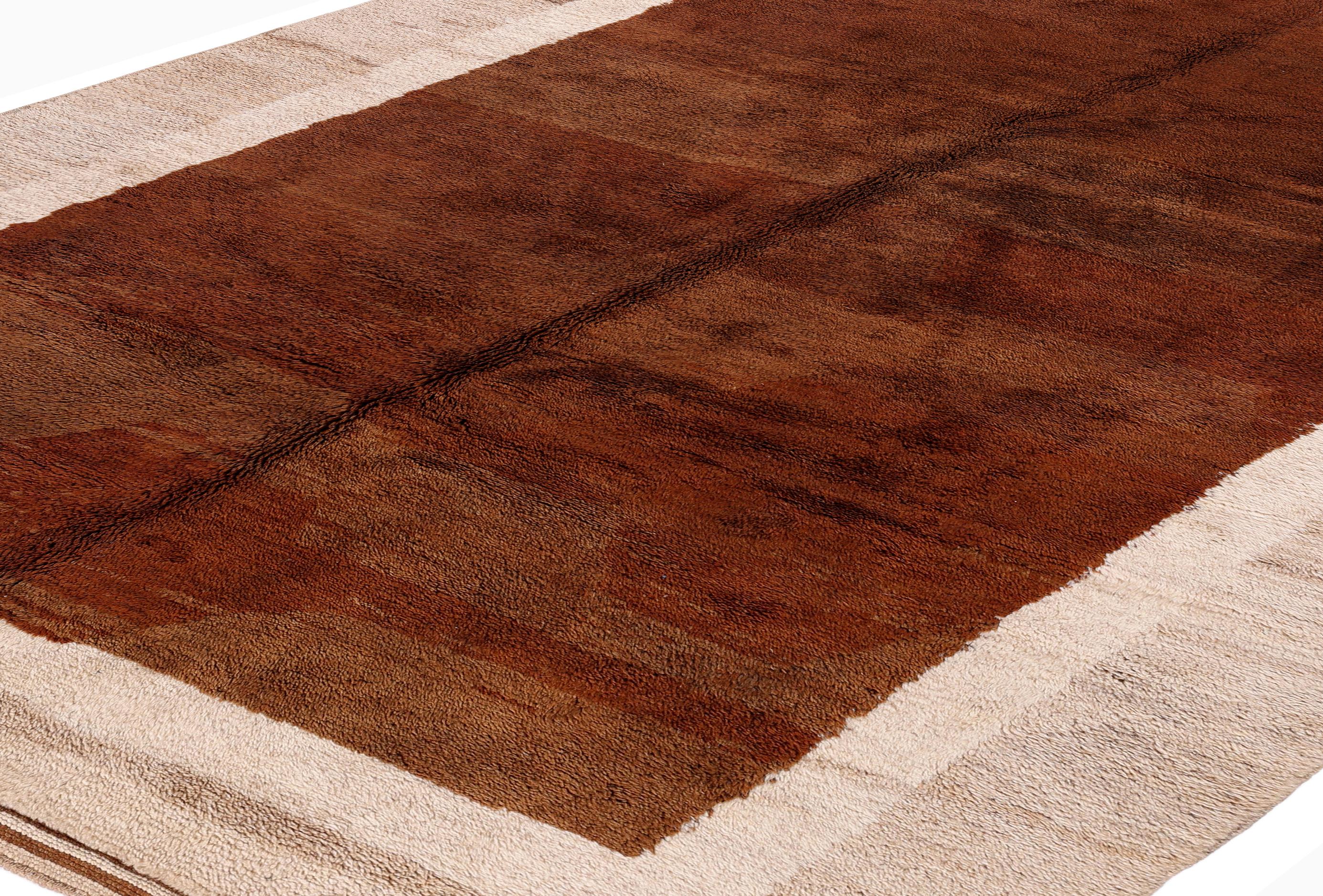 Esta alfombra marroquí única en su género está anudada a mano en lana de pelo alto lujosamente suave, y presenta un rico campo siena quemado enmarcado por un sutil borde marfil. El diseño minimalista en bloques de color combina tonos terrosos con