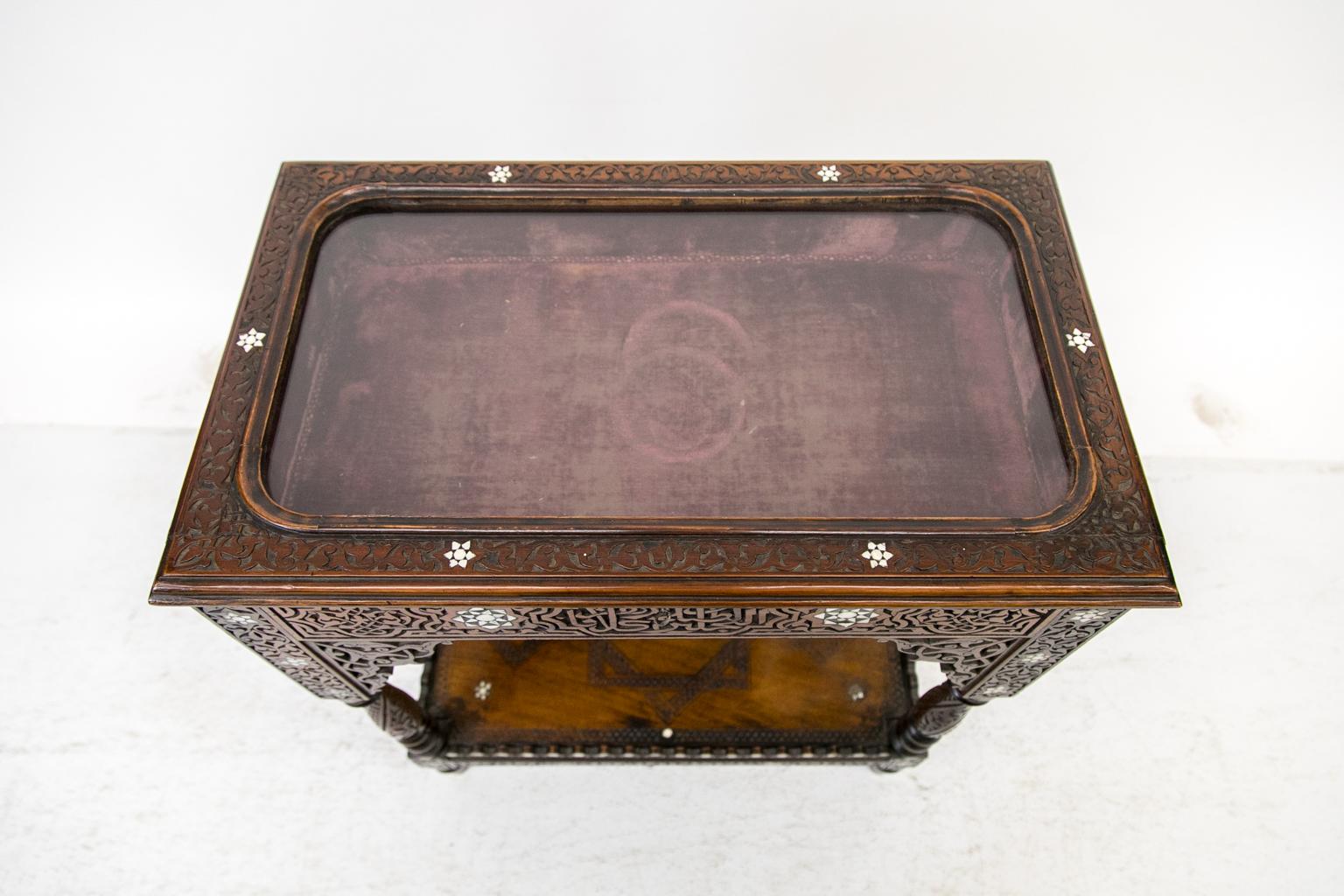 This vitrine table is carved overall with arabesques and geometric designs, and is inlaid with mother of pearl and stylized flowers. The interior display area is lined with padded burgundy baize fabric. There is a lower shelf that is carved and