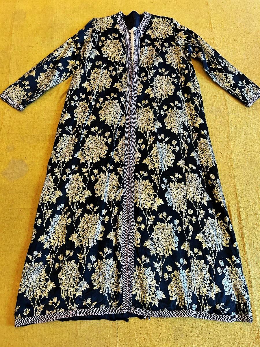 Circa 1950-1960 
Morocco 
Beautiful Moroccan ceremonial Kaftan in black satin brocaded with golden blades with a design of chrysanthemum flowers. Ample coat with bell buttons embroidered with the Jewish star and pale pink and black floche silk