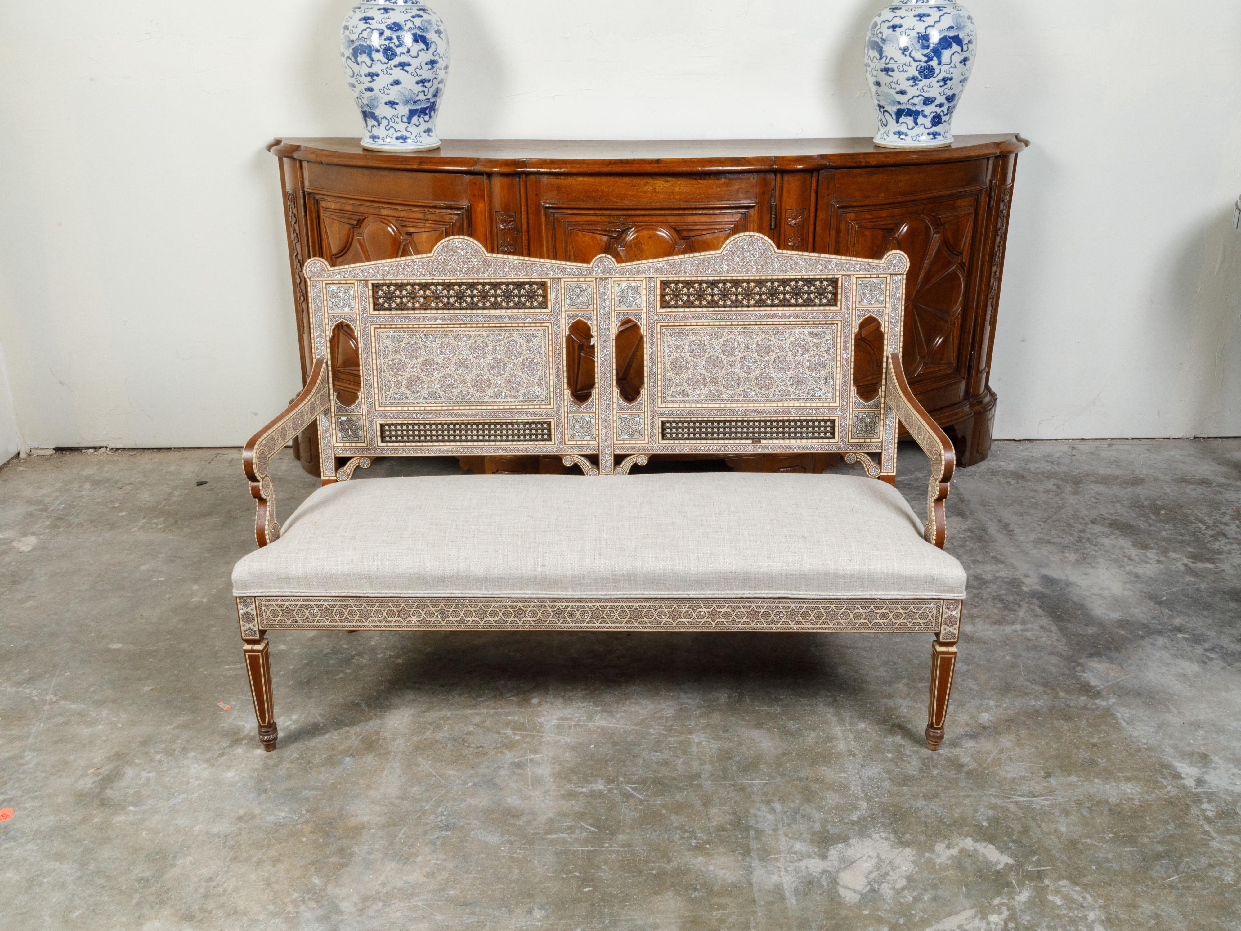 A Moroccan settee from the mid 20th century, with geometric mother-of-pearl inlay and new linen upholstery. Created in Morocco during the Midcentury period, this settee captures our attention with its stunning back adorned with an abundant décor of