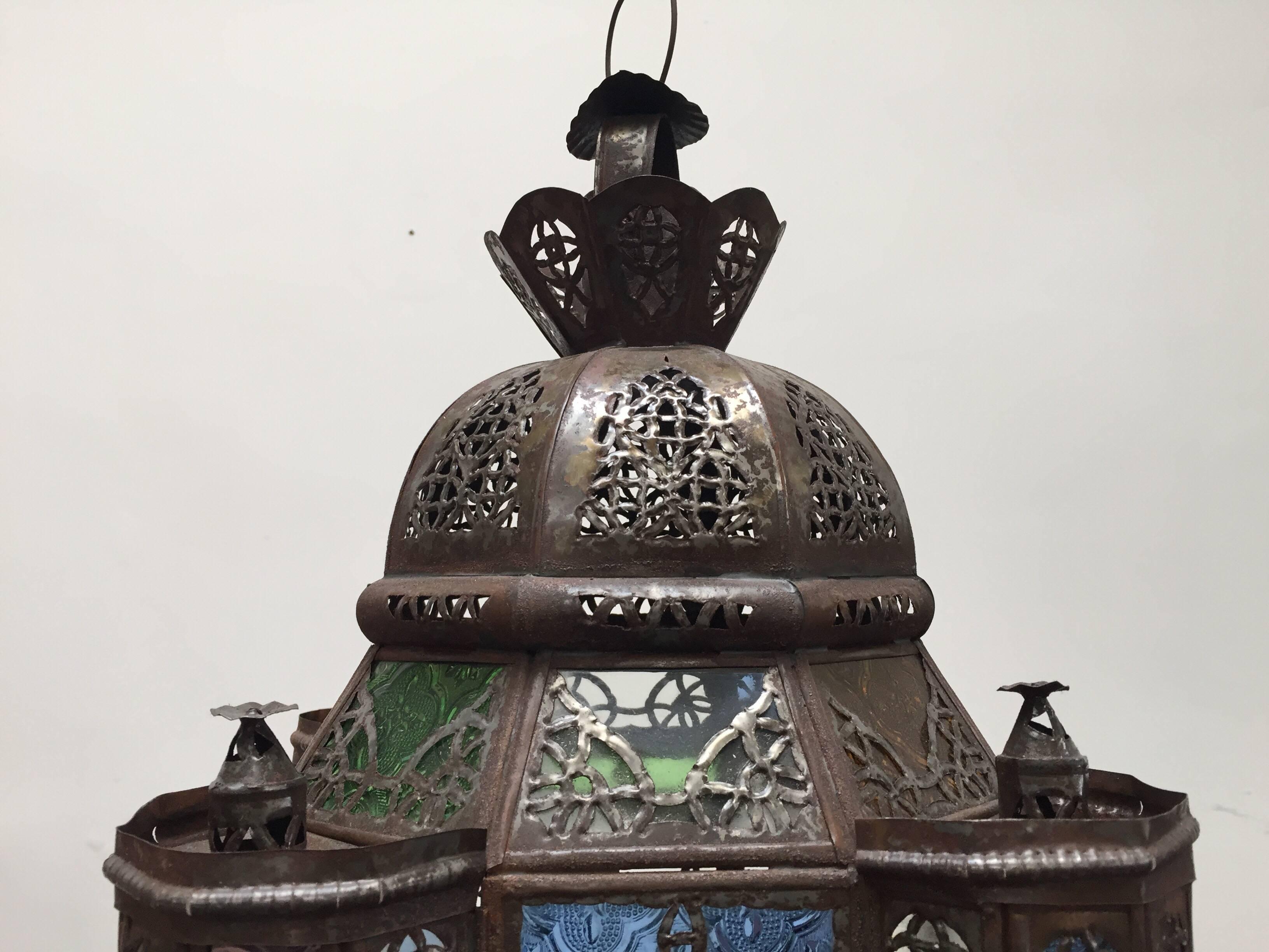 Handcrafted Moroccan glass pendant with clear and colored glass, square shape with towers and a dome with metal filigree.
This multi-color Moroccan glass lantern with Moorish arches in metal filigree and colored glass in green, blue, lavender and