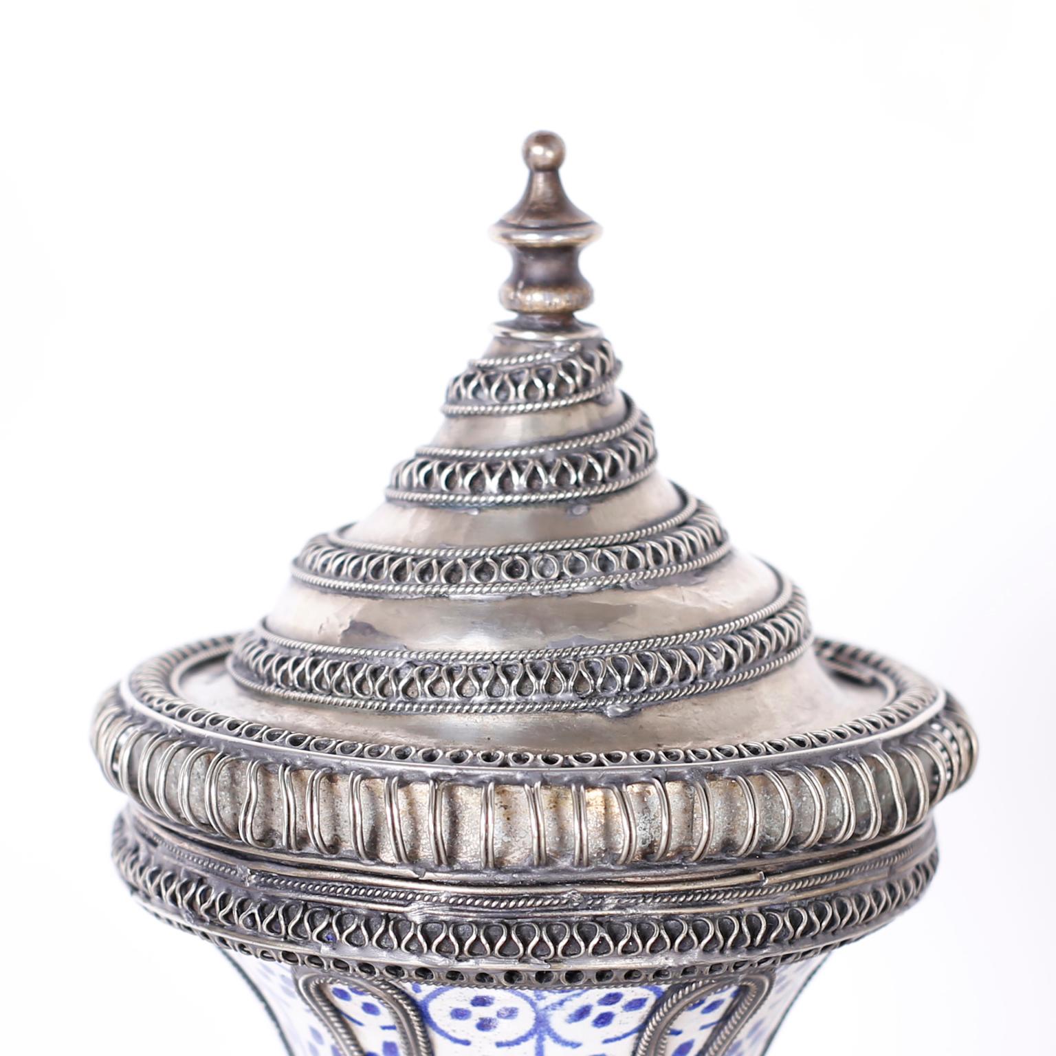 Lofty Moroccan porcelain lidded jar decorated with distinctive mediterranean designs and colors and adorned with jewelry like metal work.