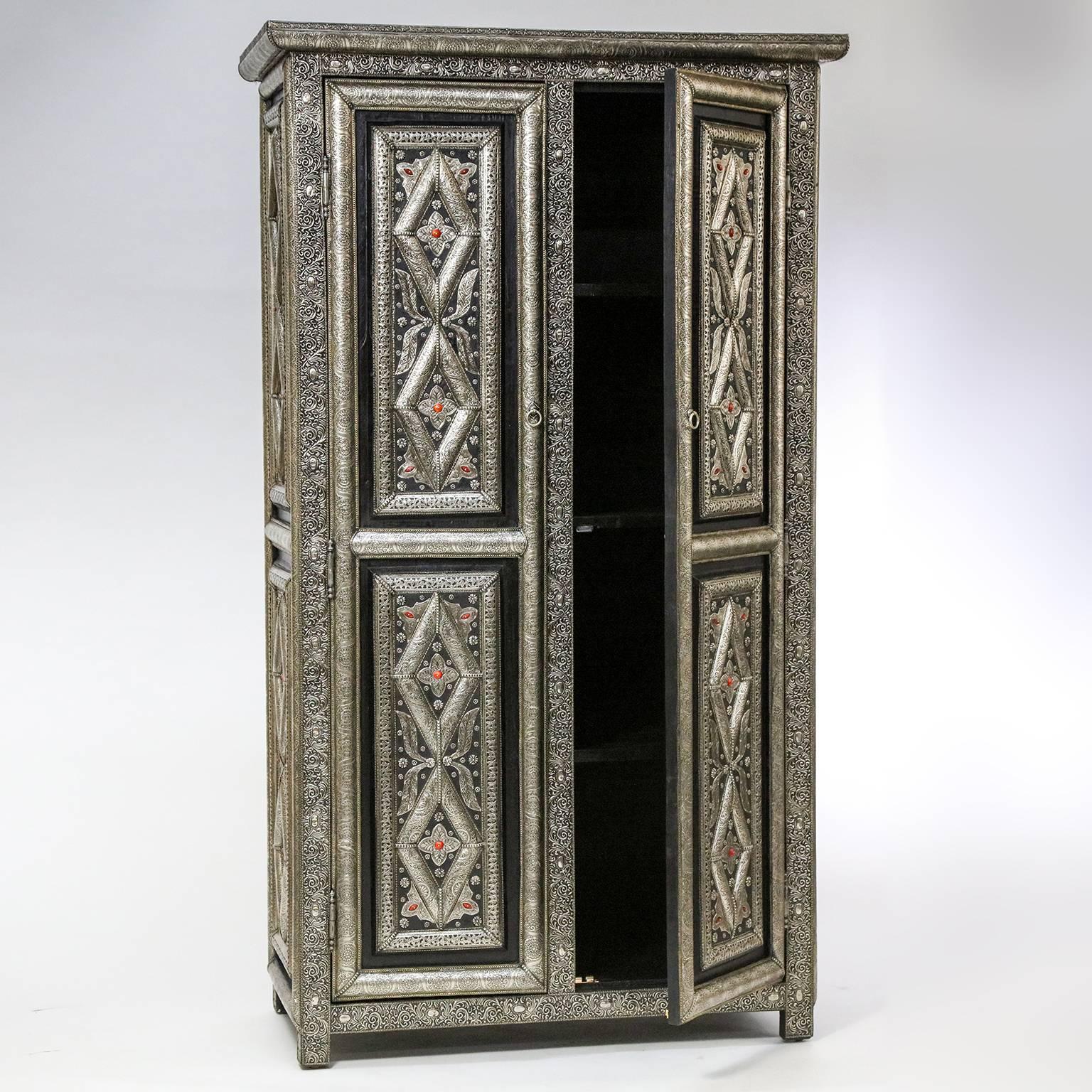 Distinctive Moroccan armoire features paneled double doors and sides, decorated with hammered and incised Moroccan silver overlay in traditional geometric patterns. Compartmentalized interior includes a clothes bar and three shelves. Each shelf is