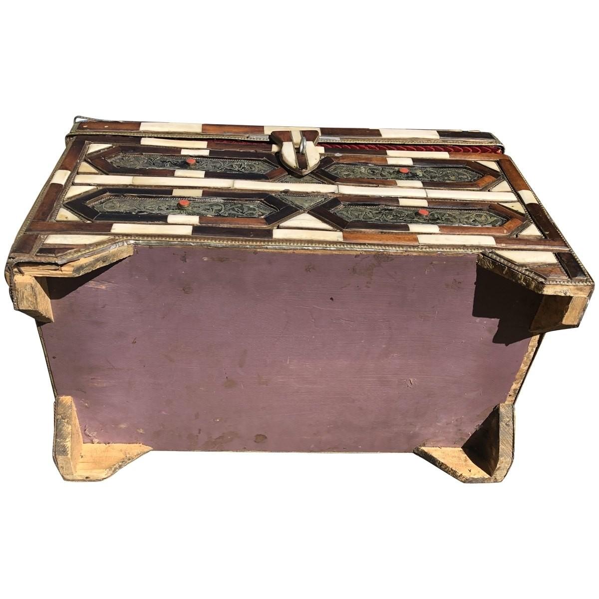 Moroccan art and furniture is defined by ornate decorations and colorful detailing. This Moroccan dowry trunk is made with a hinged lid overlaid with bone and wood. It features stamped and rope metal accents, as well as a red velvet lined
