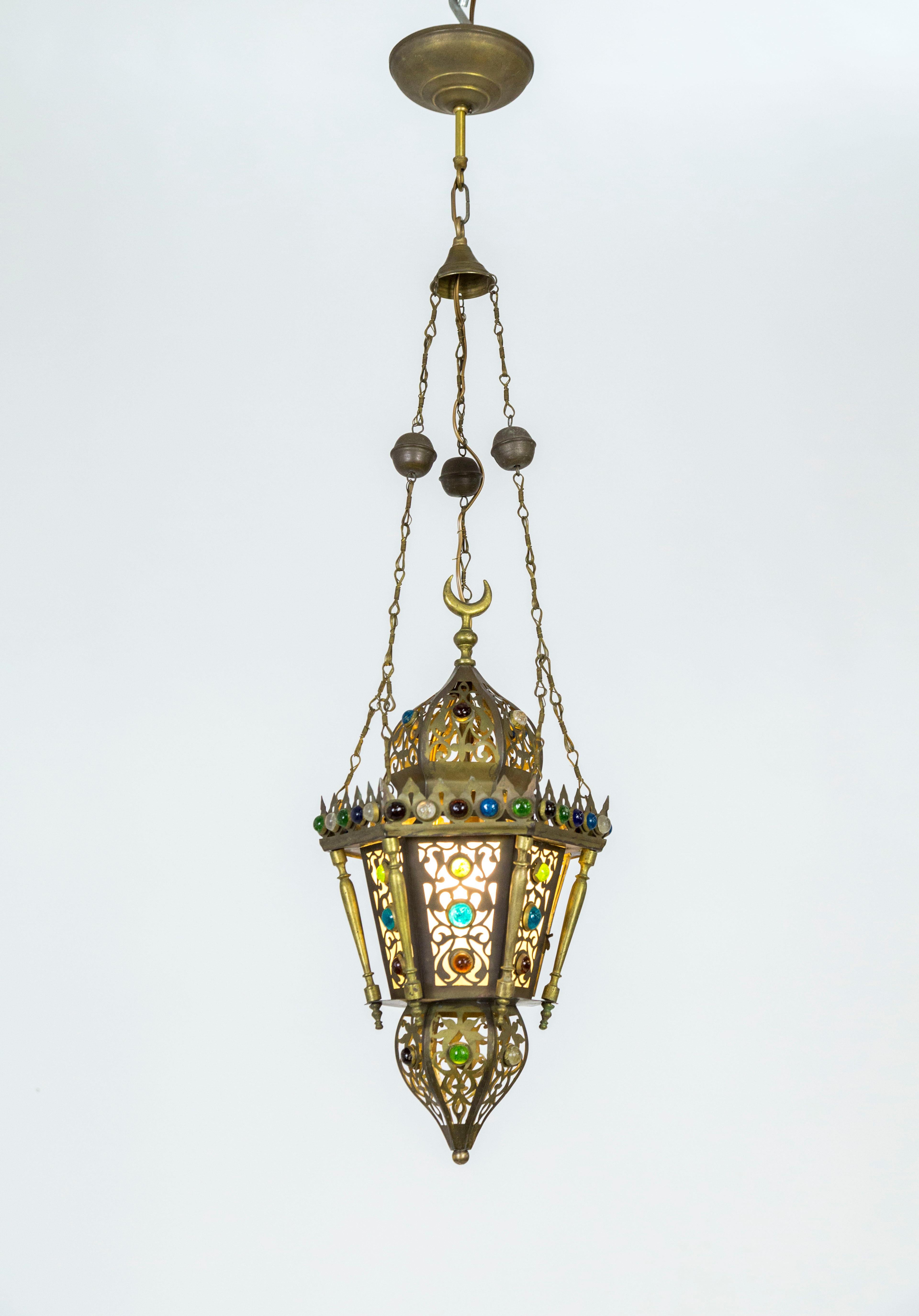 A beautifully shaped, 6-sided, brass lantern with finely cut decorations, balustrade accents, and dome top, capped with a moon shape finial; lined with blue, green, red glass jewels. Hanging from three detailed chains. Moroccan, 1930. Holds one