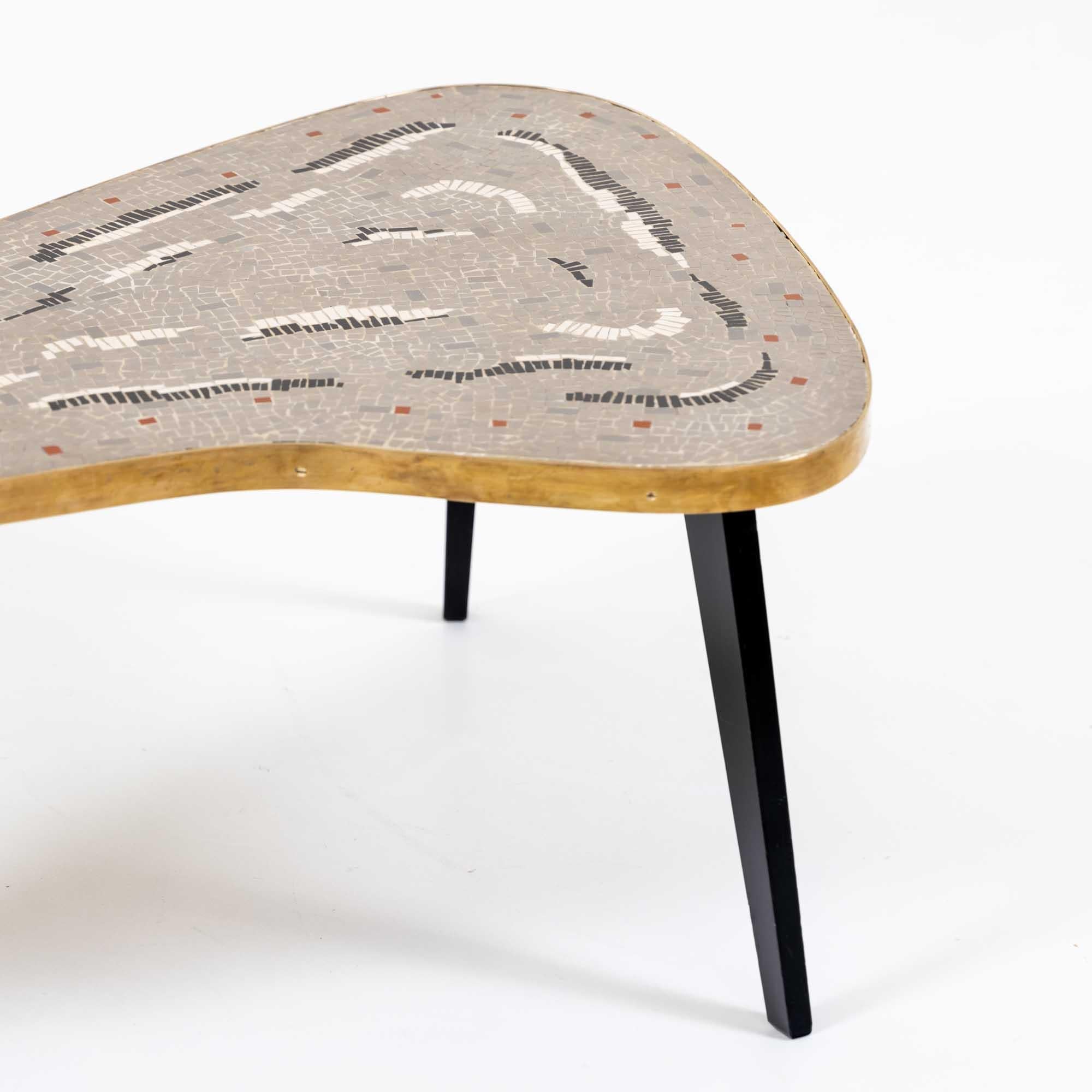 Couchtisch aus den 1960er Jahren auf drei konischen, ebonisierten Beinen. Die interessante Tischplatte ist in Form eines Bumerangs gestaltet und mit einem breiten, flachen Messingstreifen eingefasst. Das Highlight ist die graue Mosaikplatte, die mit