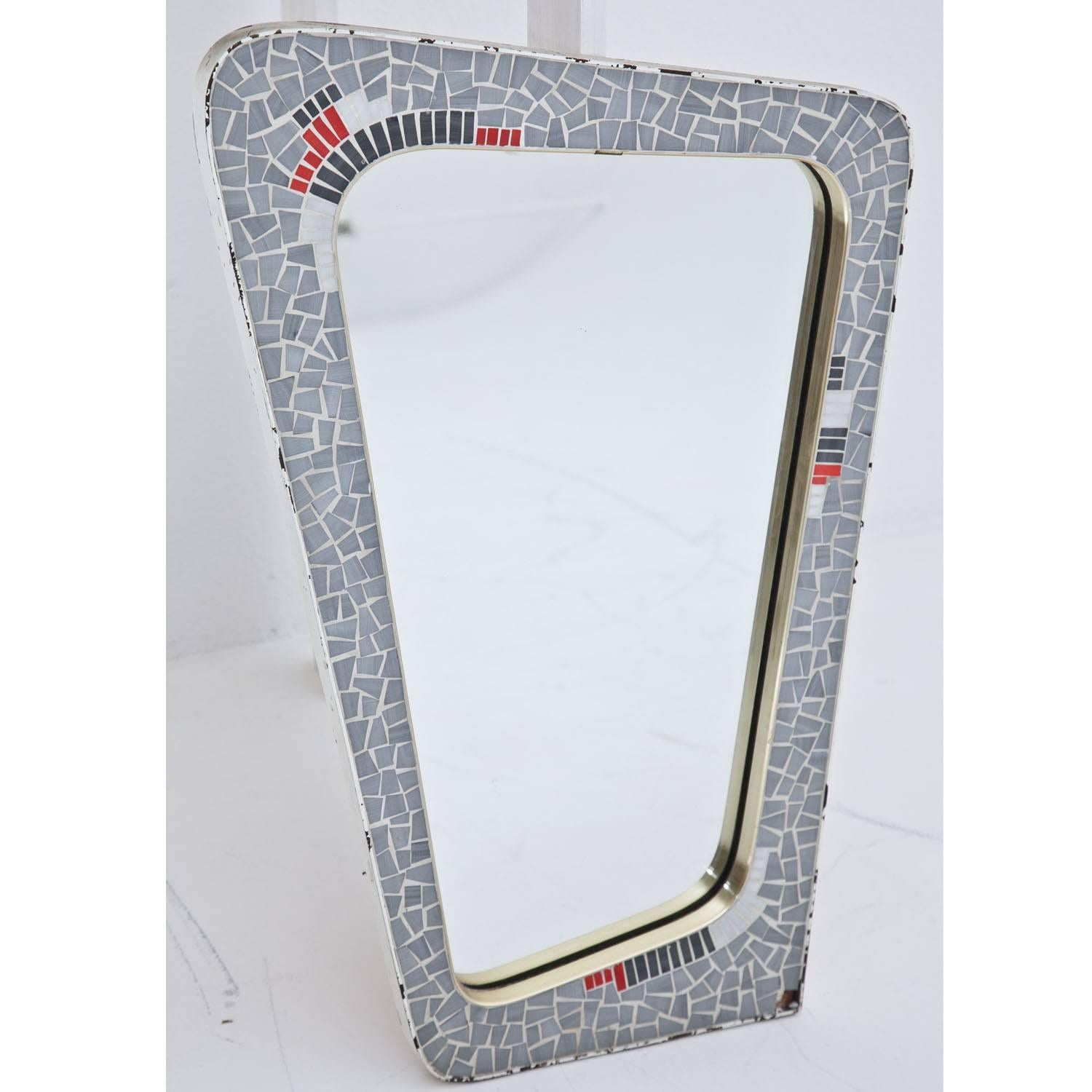 Asymmetrical wall mirror in a trapezoidal mosaic frame with blue-grey stones and red accents. One stone is missing in the bottom right corner. Label on the back, saying “Lilienthal Mosaic mirror, Model ‘Carmen’ ”.