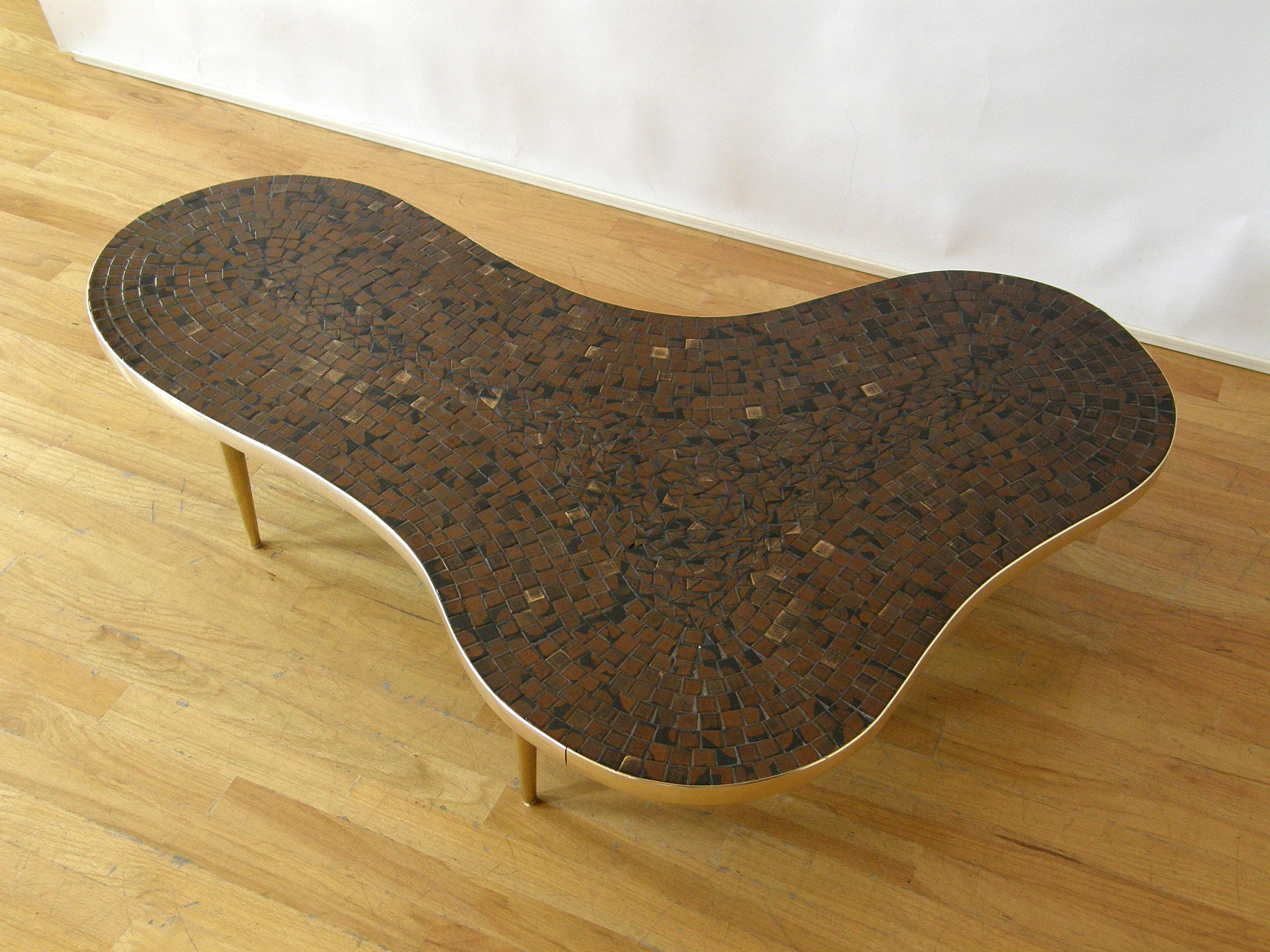 This freeform coffee or cocktail table has a mosaic tile top in shades of brown and black with scattered metallic gold accents. The amoeba shaped surface is trimmed with a gold tone anodized aluminum edge, and it rests on four tapering dowel legs in