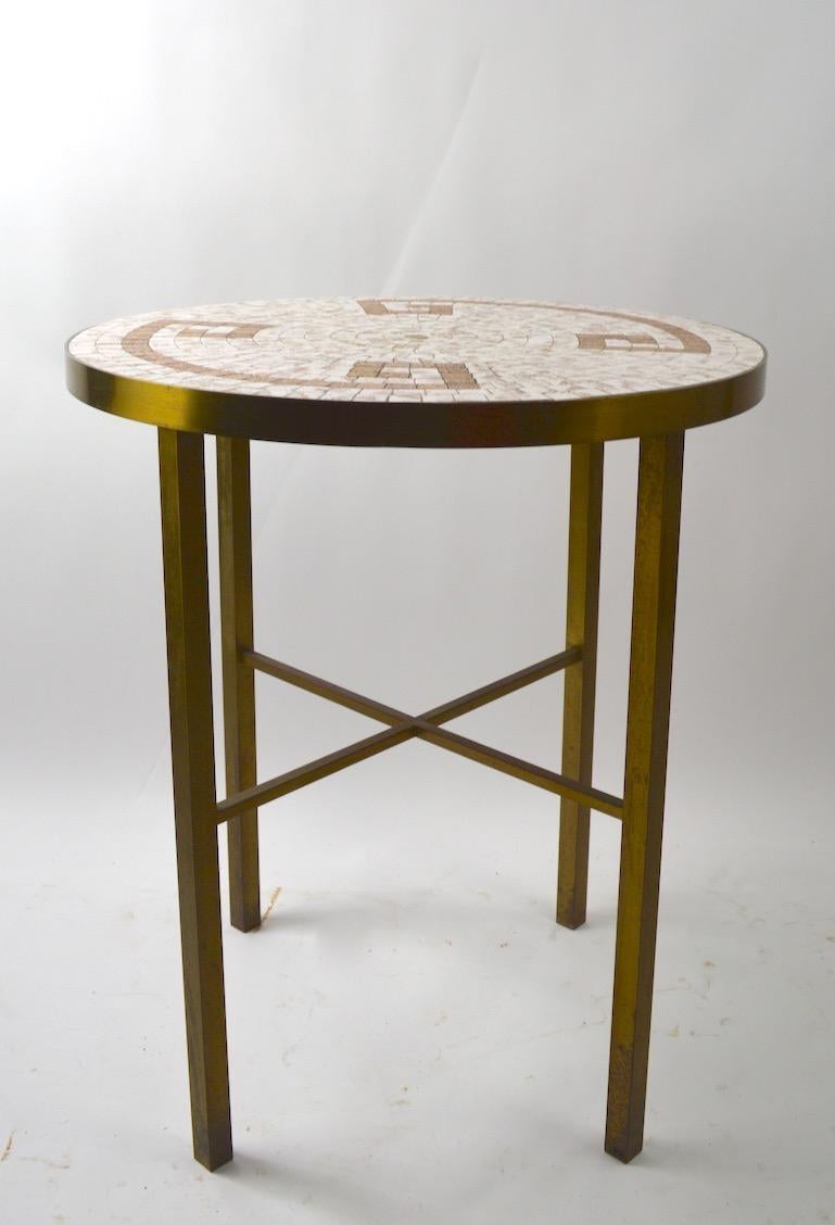 Circular mosaic tile top table with squared brass legs and stretcher. The top has a Roman Key motif in brass tone tile with metallic and off white tile ground.