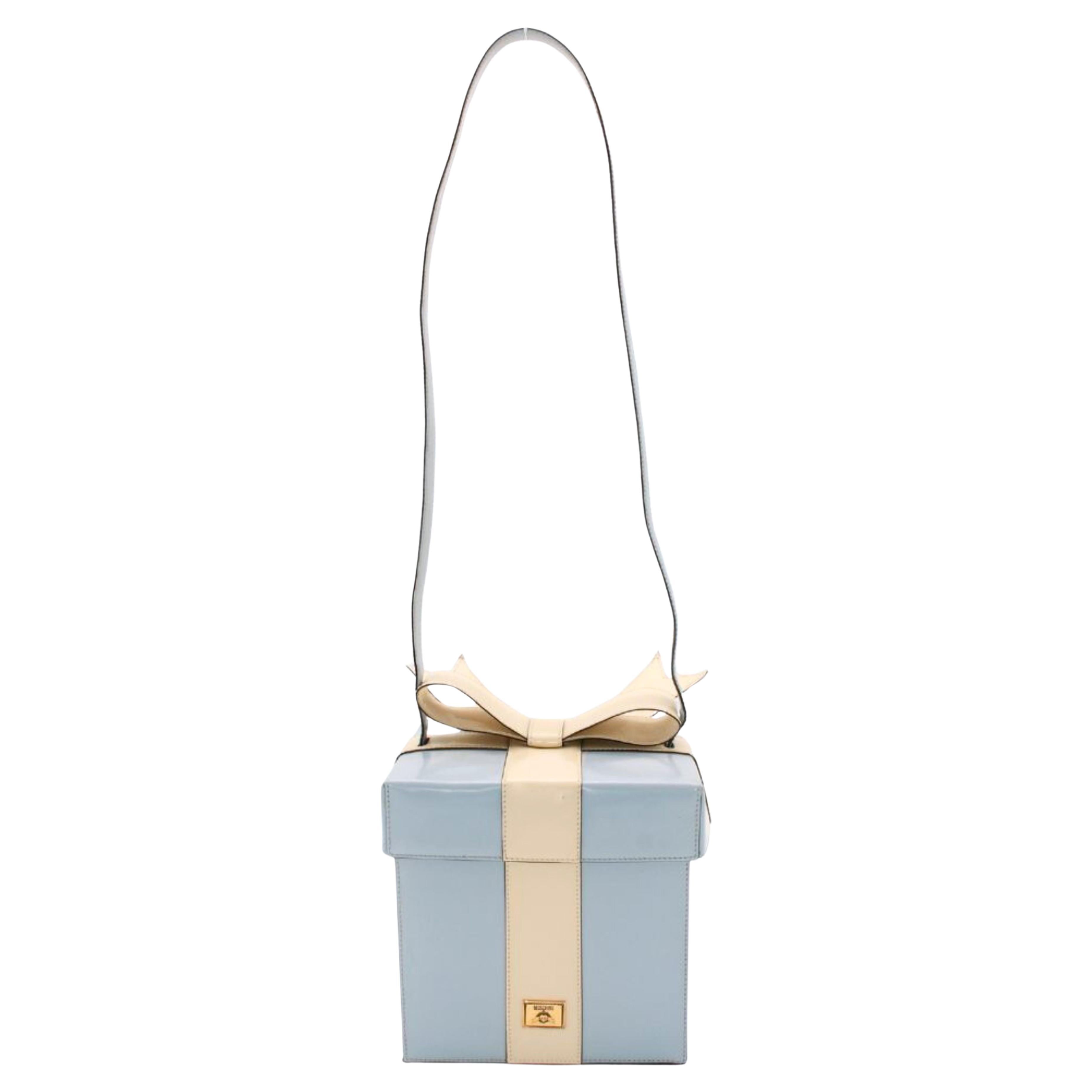 Datant des années 90, cet incroyable sac Moschino s'inspire d'un coffret cadeau de Tiffany. Confectionné en cuir verni bleu et crème brillant, il présente un nœud sur le dessus, une bandoulière, une plaque d'authenticité Moschino dorée et une