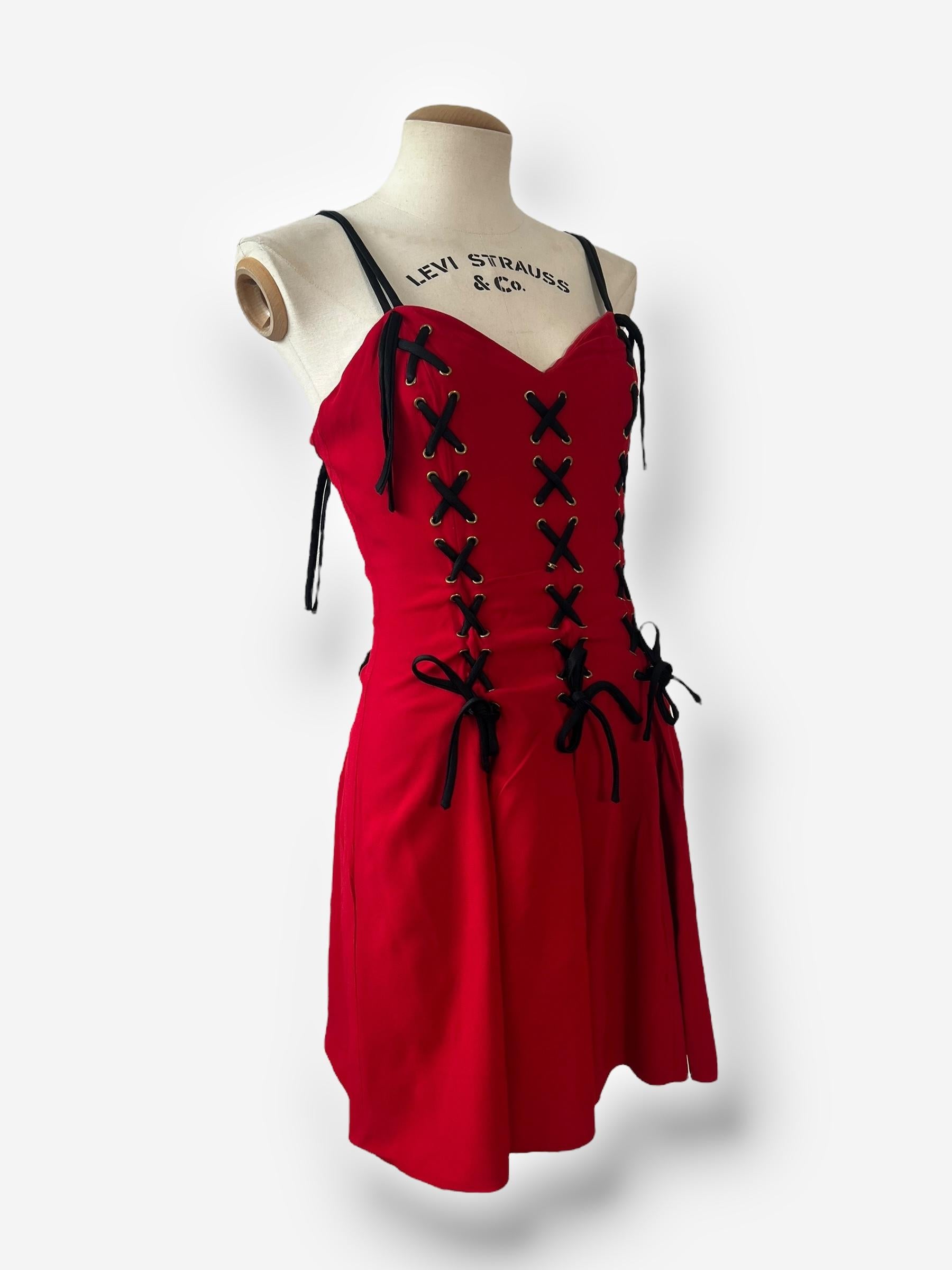 

Un pezzo iconico di Moschino Cheap and Chic, questo vibrante mini abito rosso cattura il romanticismo sfacciato e l'irriverenza giocosa per cui il marchio è famoso. Caratterizzato da audaci dettagli in nastro nero allacciato sul davanti e sui