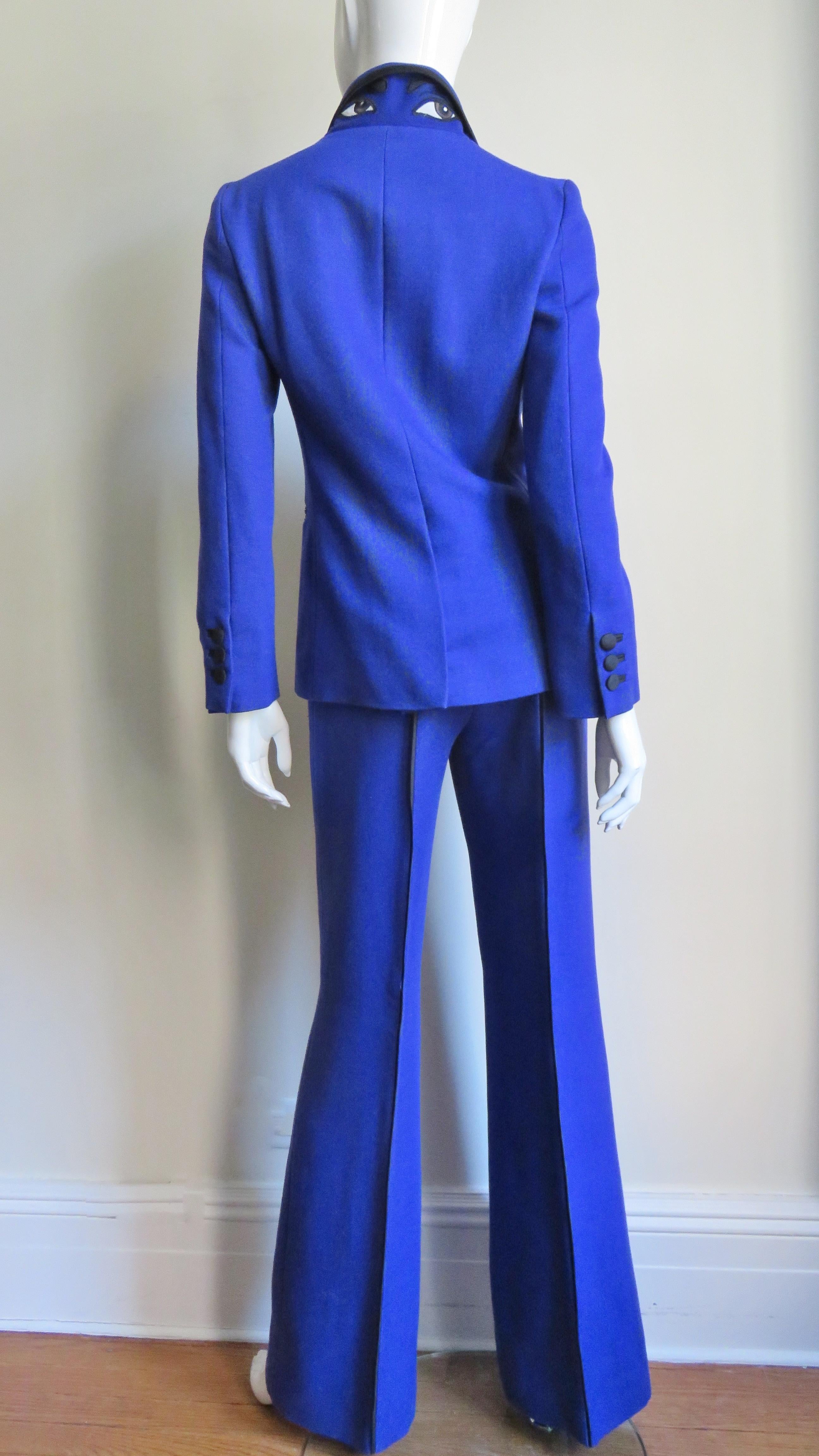 A fabulous bright blue wool pant and jacket suit from Moschino with detailed embroidered appliques of eyes and eyebrows on the back collar.  The jacket has a lapel collar,  breast pocket, front black button closing, button cuffs and hip pockets all