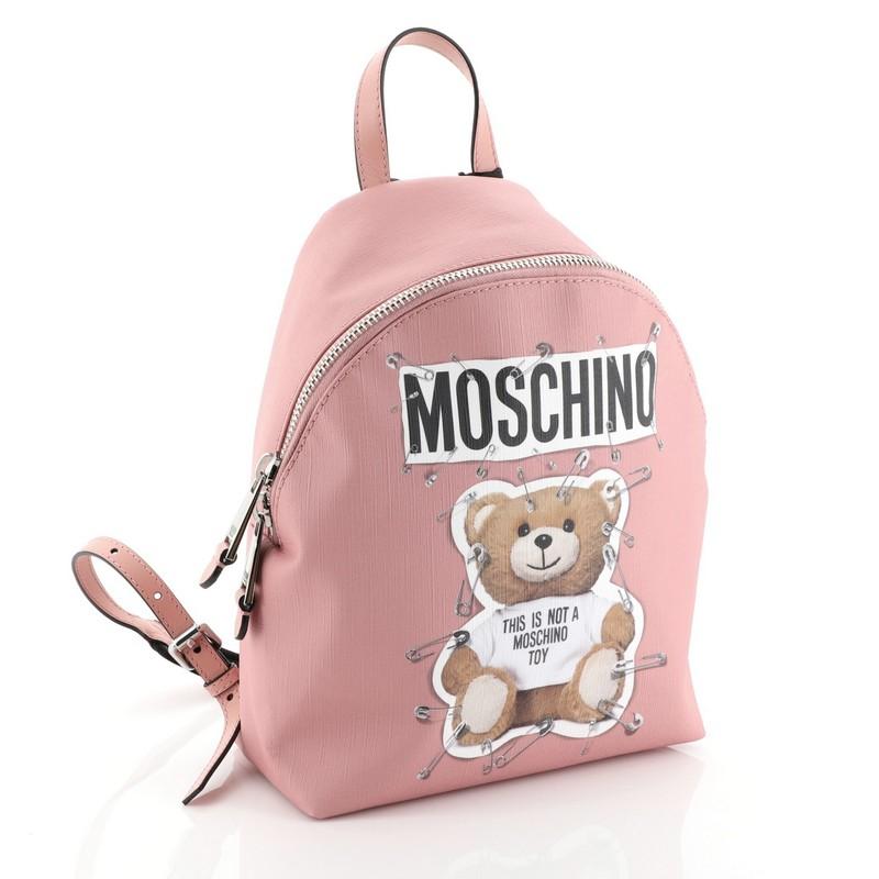 This Moschino Teddy Bear Backpack Printed Leather, crafted in pink printed leather, features adjustable backpack straps, teddy bear print, front zip pocket, and silver-tone hardware. Its top zip closure opens to a black fabric interior with zip and