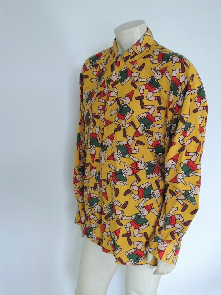 Vintage Moschino Jeans button-down shirt with a vibrant Pinnochio print. Yellow base with red, green and tan Pinnochio figures. Made in the USA, 100% cotton corduroy. 

The shirt is in fair vintage condition. This is a used, vintage garment that has