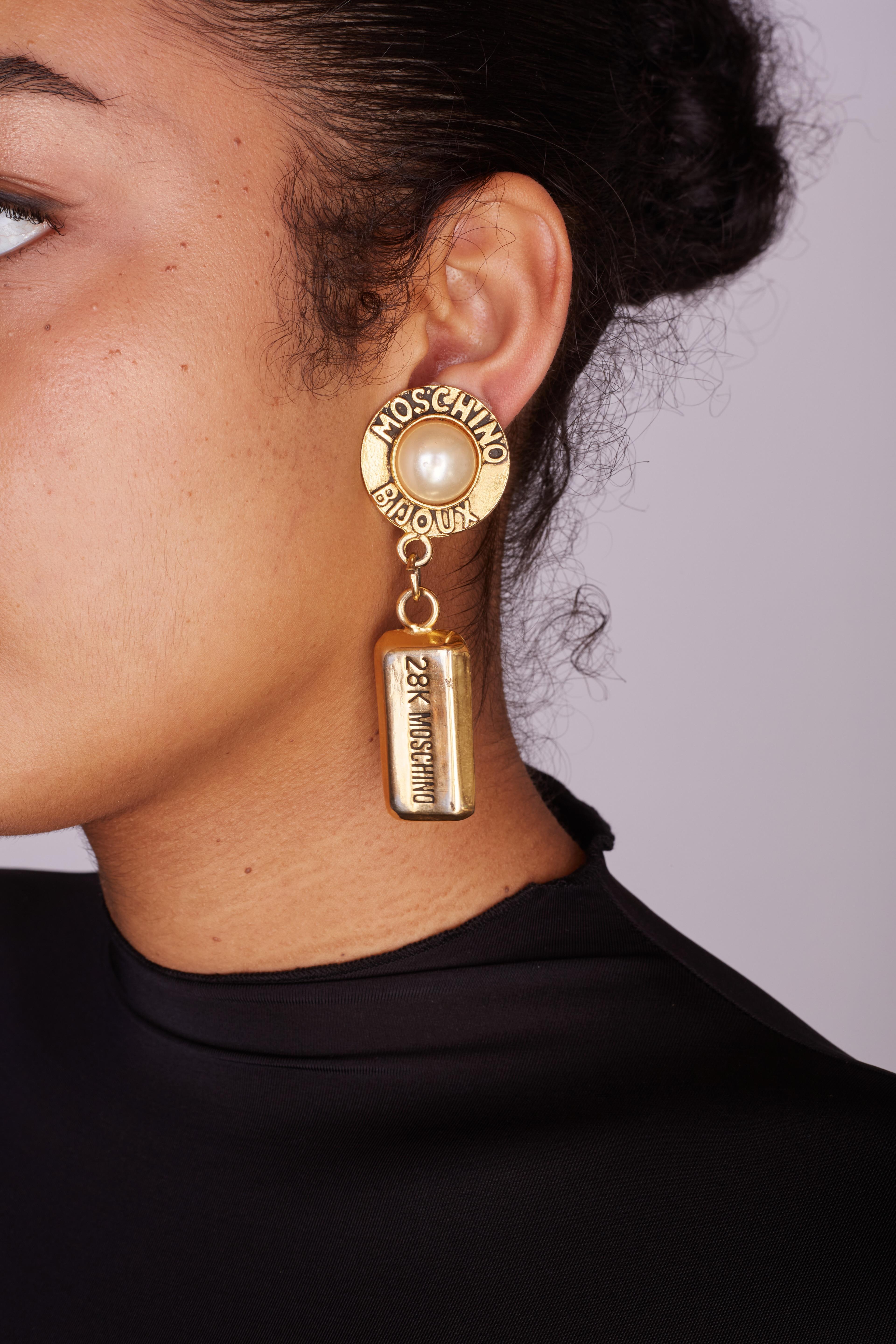 MOSCHINO VINTAGE CHUNKY GOLD BRICK PEARL EARRINGS

The vintage Moschino drop earrings from the 1990s feature a large faux pearl bead cabochon, a large metal bar motif pendant drop, gold tone color and are clip on earrings. Moschino Bijoux and 28K