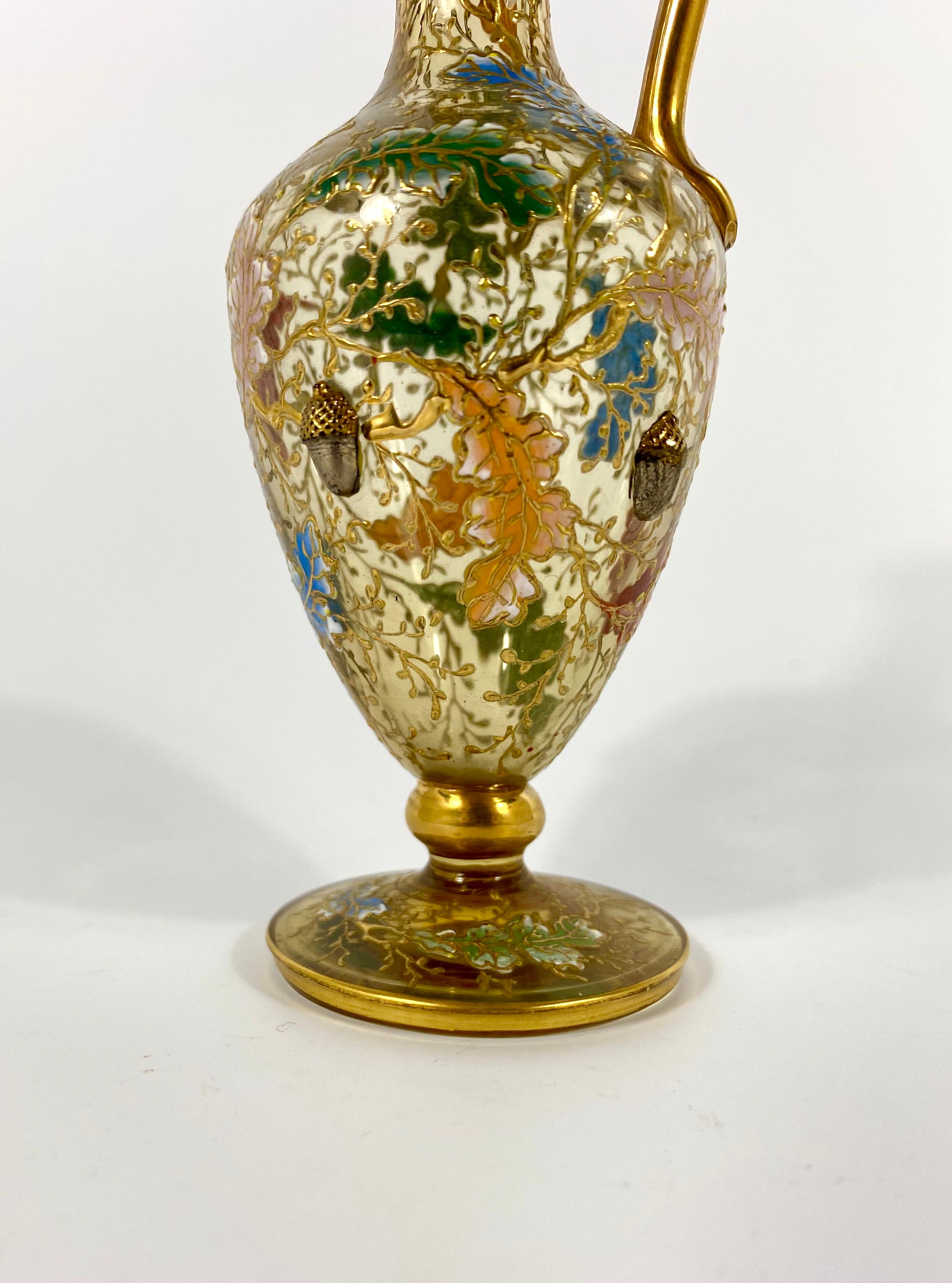 A Moser, miniature enameled glass ewer, circa 1890. The elegant classical shaped ewer, moulded with acorns, hanging amongst enameled leafs, from gilt branches, with a large insect flying above. Having a simple gilt strap handle, and set upon a