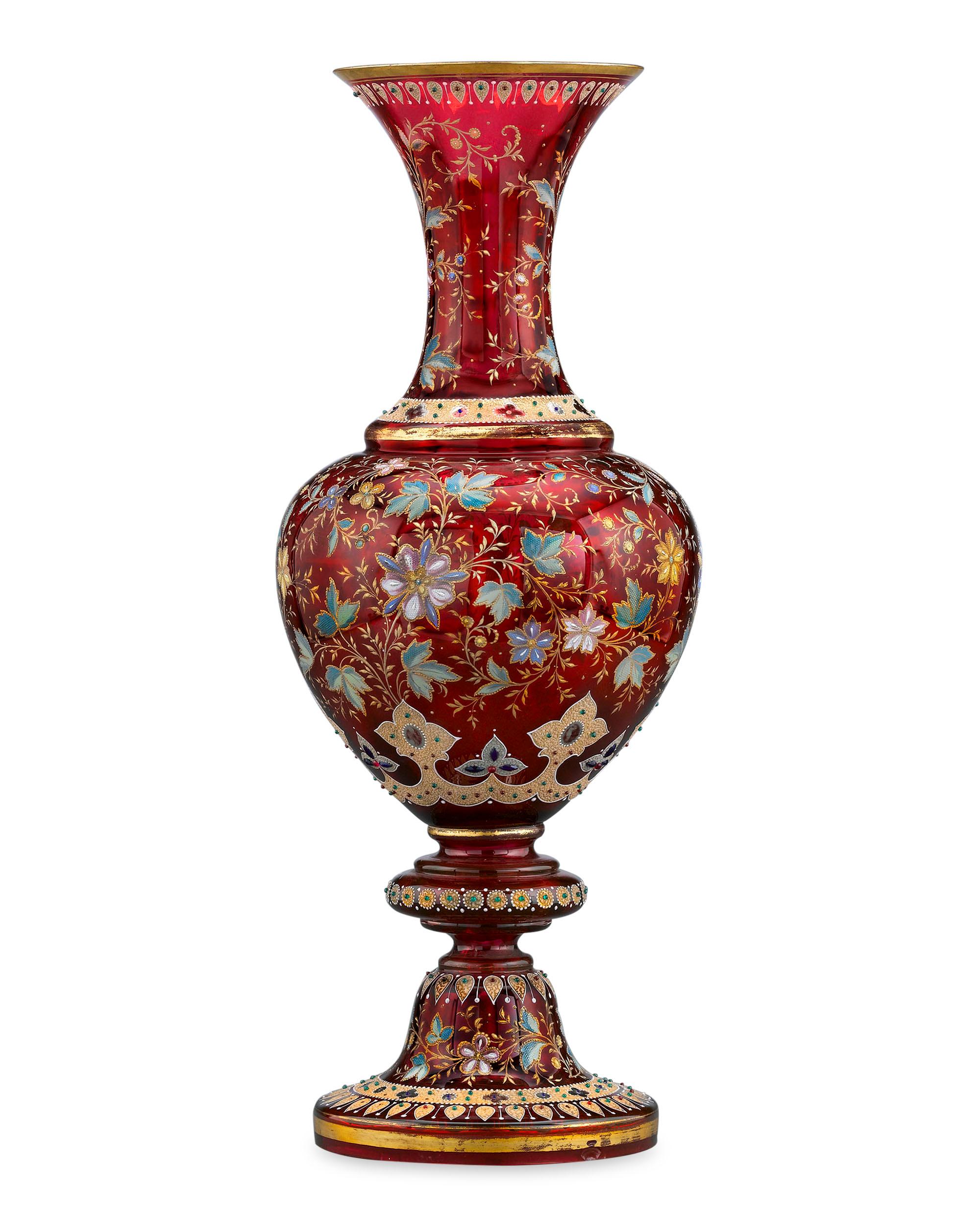 Almost certainly made as an exhibition piece, this monumental Moser ruby glass vase boasts a gilt-accented design of textured oak leaves and flowers, all enclosed by textured bands of jeweled gilding. Formed in a classical urn shape in Moser’s