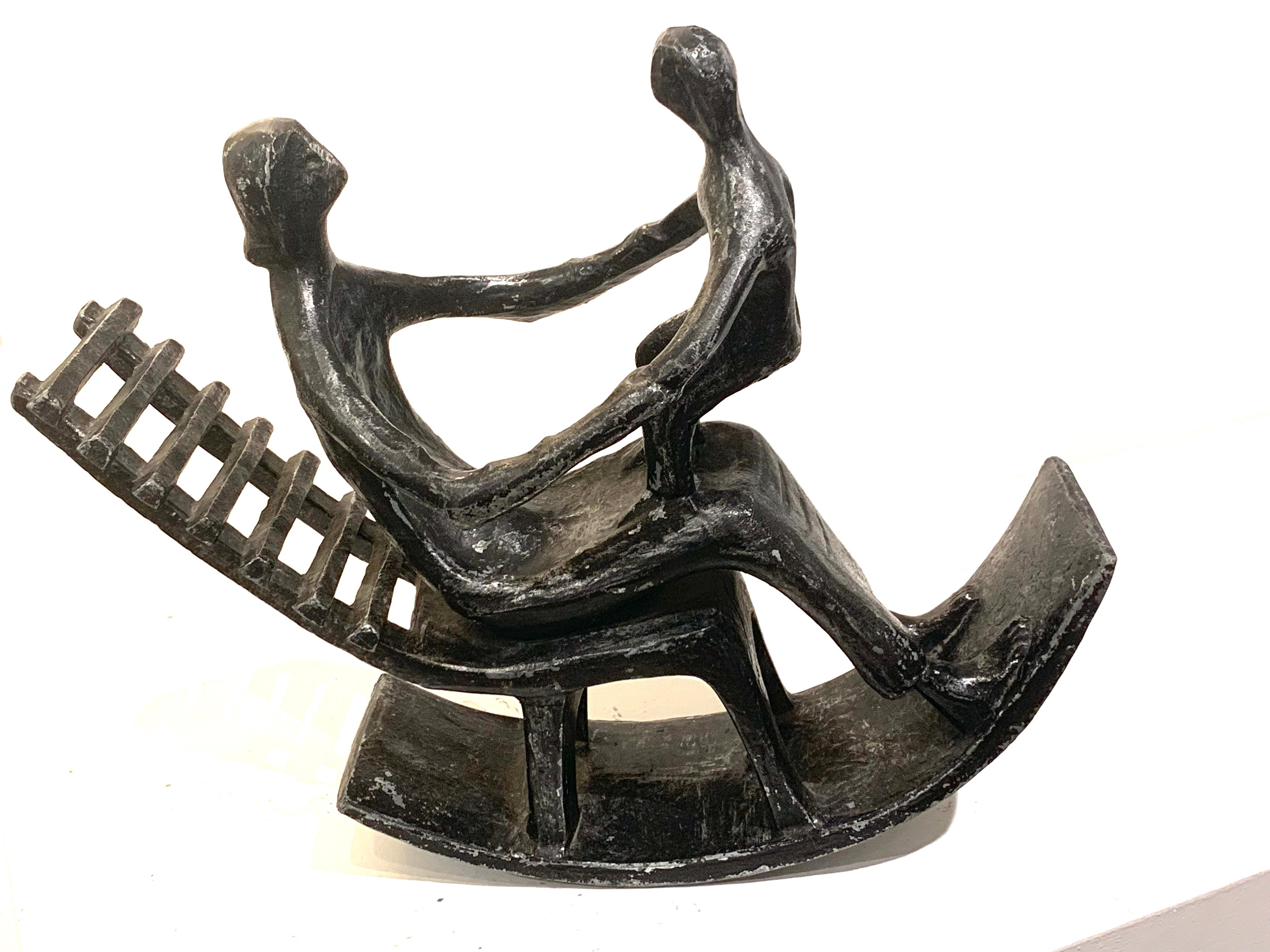 Scultura in metallo brunito di madre e figlio che si godono un momento di gioco su una sedia a dondolo. La prima immagine mostra la scultura in equilibrio naturale sul fianco della madre. L'immagine 3 viene tenuta per aumentare l'inclinazione e