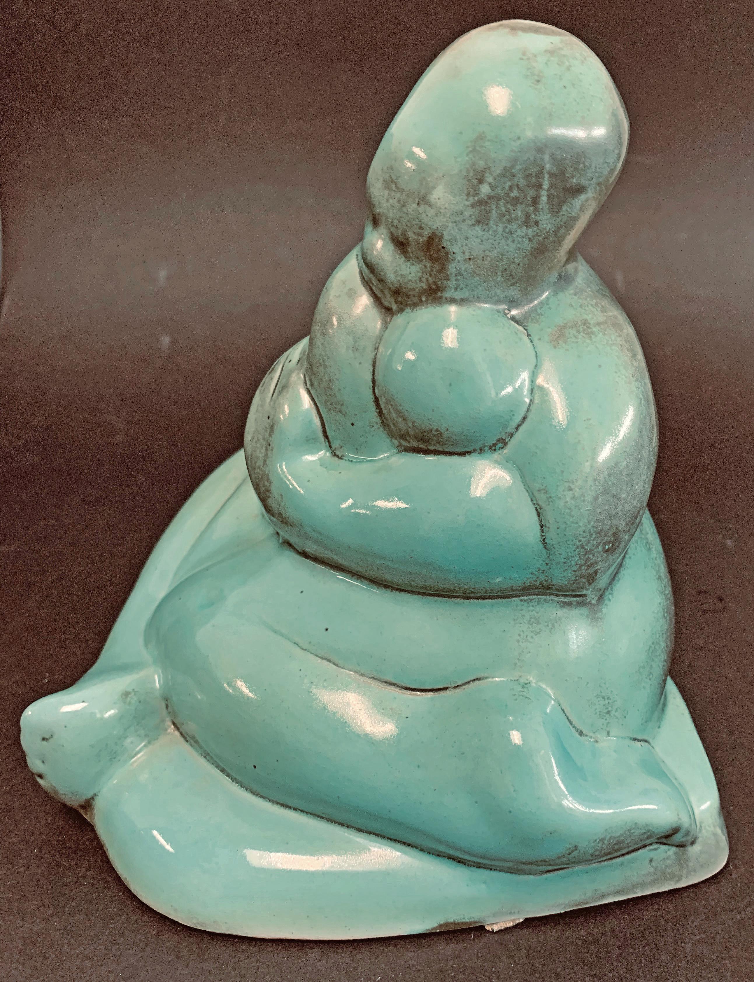 Stylized and reductive, this beautiful 1930s-vintage ceramic sculpture depicts a sitting mother holding her child very close, her legs crossed and her arms enfolding the baby. The warm turquoise color is highlighted with areas of charcoal-hued
