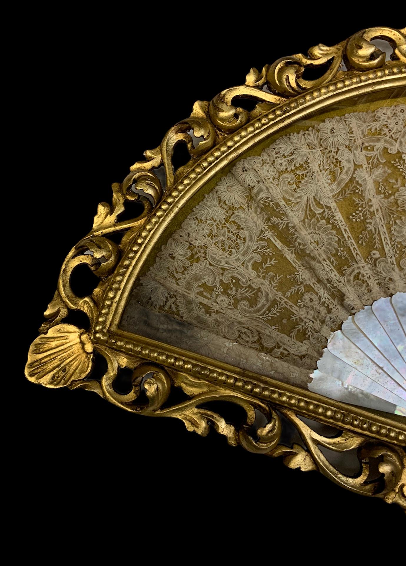 This is a delicate mother of pearl sticks and Lace fan enclosed in a fan shaped gilt wood & glass display. The border of the box is adorned by golden beading. Over this border is highlighted a carved decoration made of scrolls of acanthus leaves
