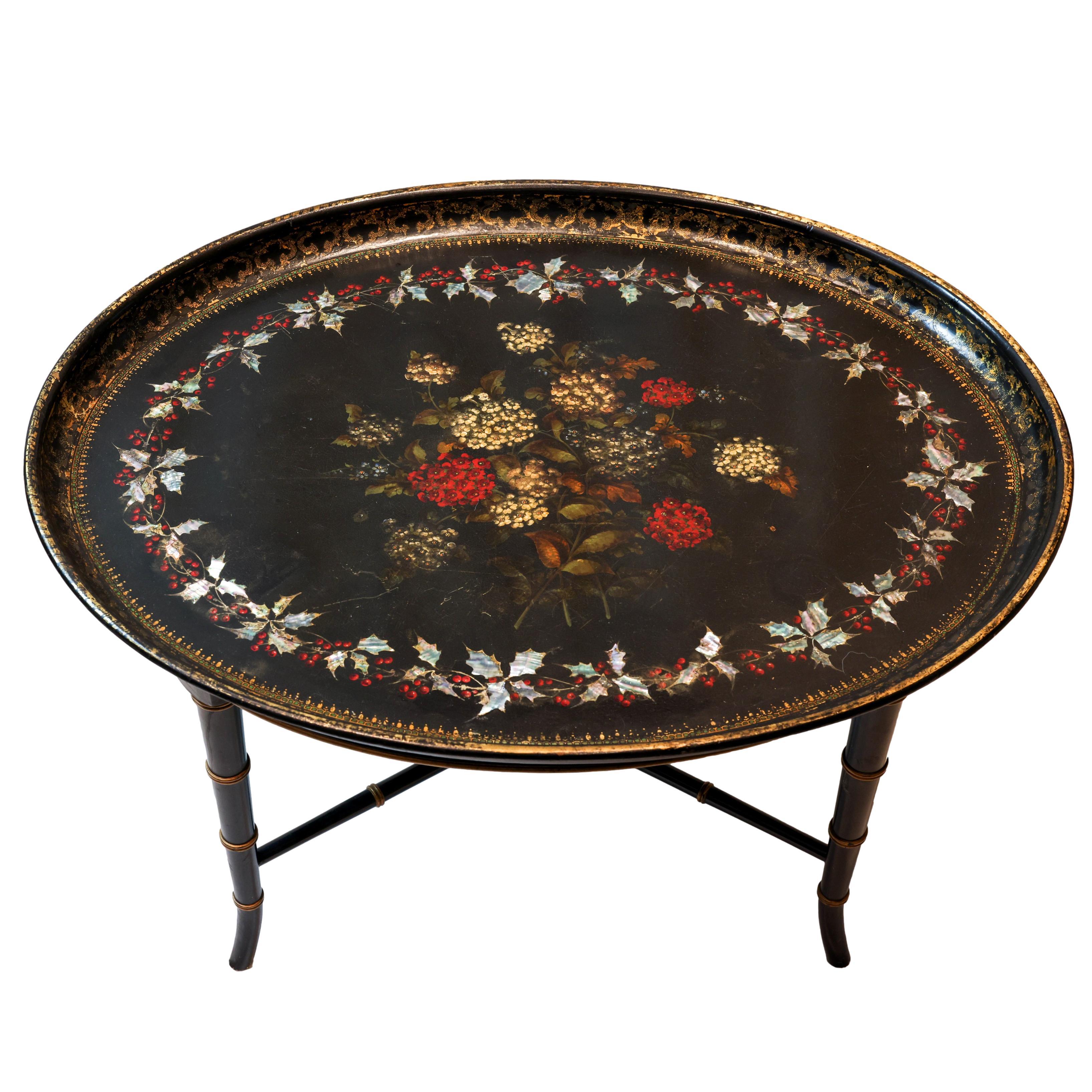 Ebonized paper mache tray table, the oval tray with a central painted still life of hydrangeas, with encircling band of inlaid mother-of-pearl holly leaves and painted red berries, with gilded borders and rim, on a later faux bamboo conforming base.