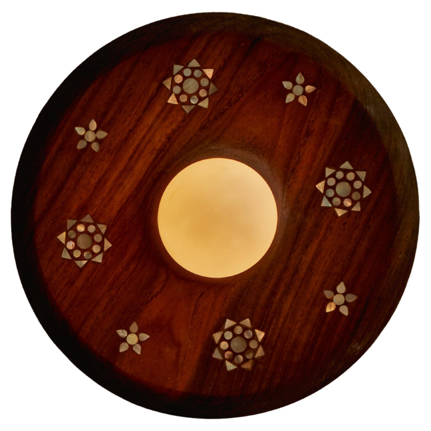 L'applique Mother est une ode à la lumière et à l'éducation. Fabriqué à partir de teck récupéré et incrusté de nacre, il équilibre la chaleur de la terre avec l'éclat céleste. La forme ronde, incrustée de délicats motifs floraux étoilés, rappelle