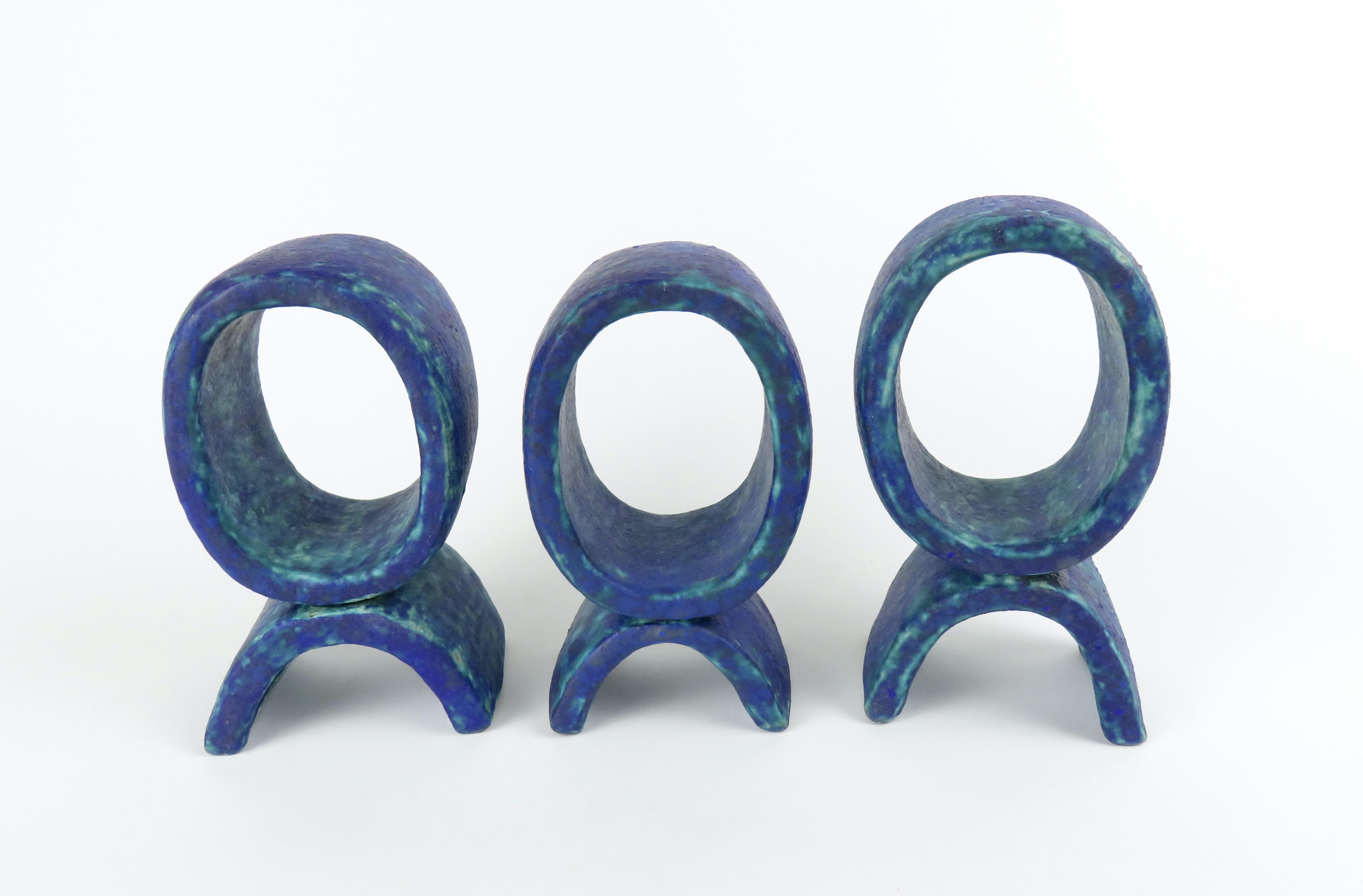 Trois petits totems en céramique de couleur turquoise et bleu foncé. Ils se composent chacun d'un seul anneau vertical sur un petit pied incurvé. Chaque pièce est construite à la main en grès céramique, attachée ensemble et cuite deux fois. Le plus
