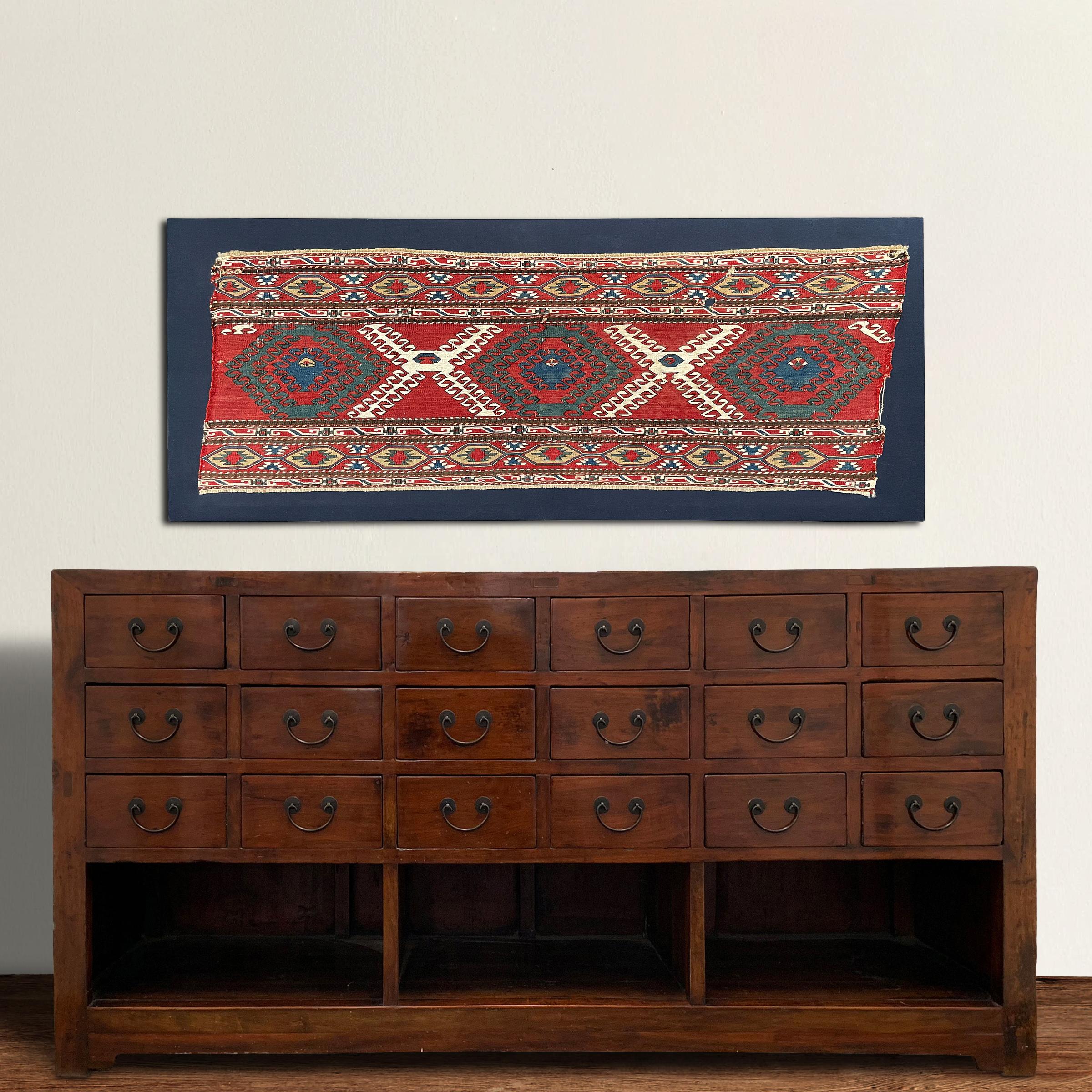 A bold mounted 19th century Azerjaibjani Soumak rug fragment containing several stylized floral patterned motifs and stripes containing petite floral and geometric patterned stripes, all woven in light and dark indigo, crimson, green, white, and