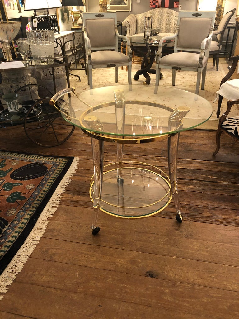 A super glamorous French gold-plated brass, Lucite and glass round bar cart having an Art Deco influence. The 3 splayed legs are incredibly elegant tapered Lucite with sculpted brass caps at the top and pretty casters that move silkily. The handle