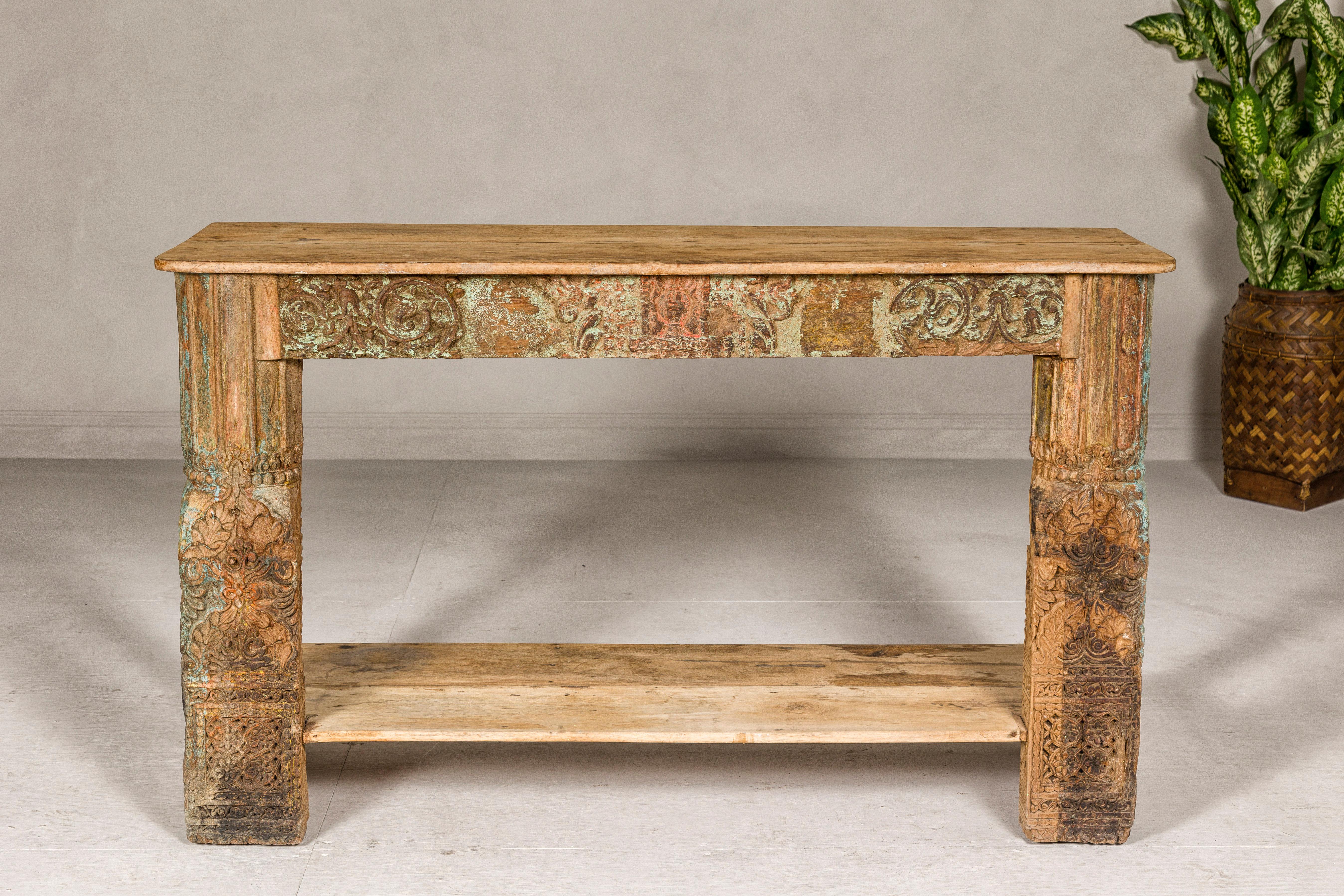 A Mughal style polychrome console table with scrollwork carved apron, foliage décor, bottom shelf and distressed appearance. Experience the splendor of Mughal artistry with this contemporary polychrome console table, a piece that beautifully