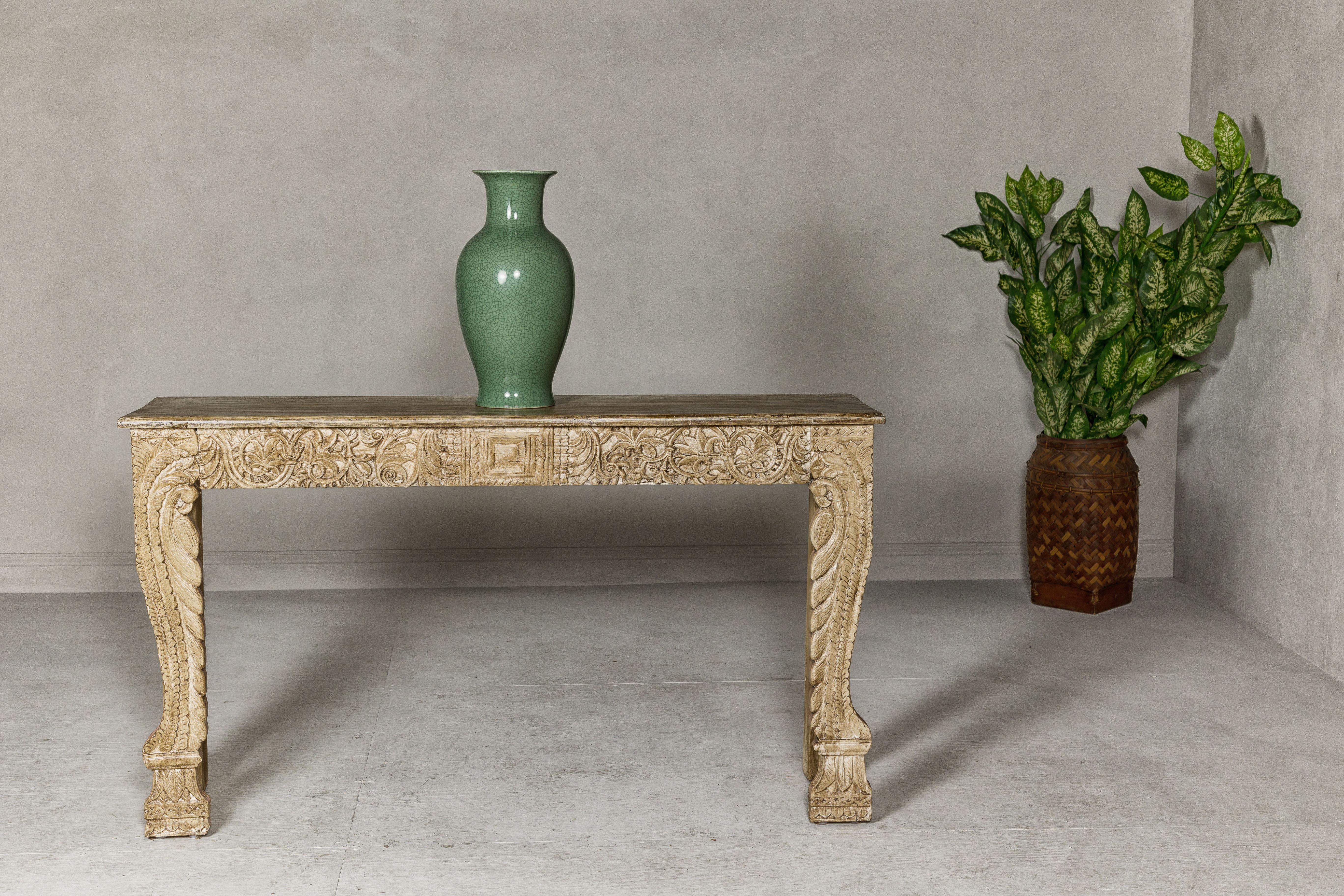 A Mughal style painted console table with carved apron and legs. This Mughal style painted console table captivates with its exquisite craftsmanship, featuring an intricately carved apron and legs adorned with abundant foliage motifs. The table