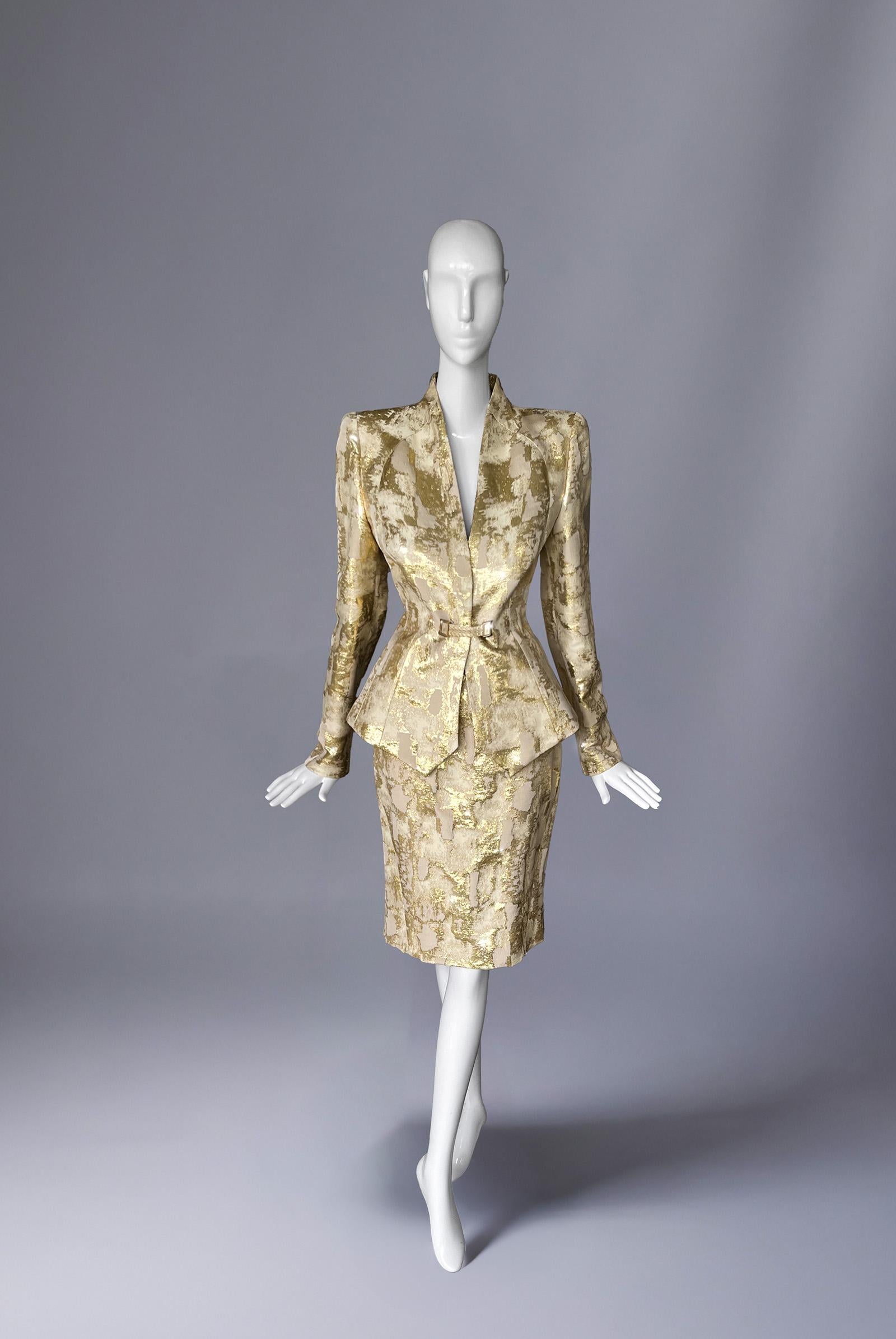 In una sinfonia di precisione scultorea e scintillio celestiale, questo ensemble Mugler emerge come una masterclass di alchimia couture. Realizzato in una sontuosa miscela di lana, seta e lamé d'oro, l'abito irradia un'opulenza imponente, un