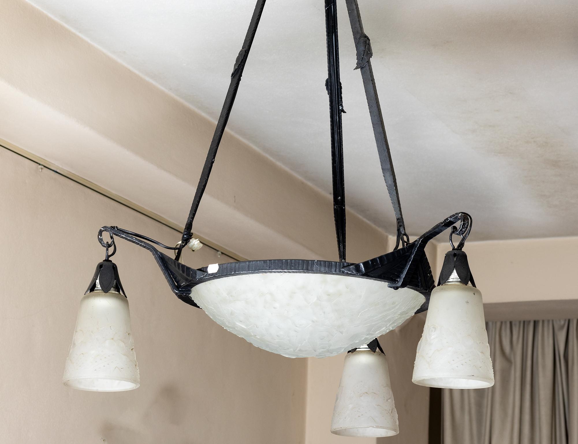 Questo lampadario Art Déco degli anni '20, firmato Muller Frères, presenta una struttura in ferro battuto scuro che sostiene una ciotola centrale in vetro smerigliato con tre paralumi a tulipano. 

Il vetro opalino sfoggia motivi geometrici Art Déco
