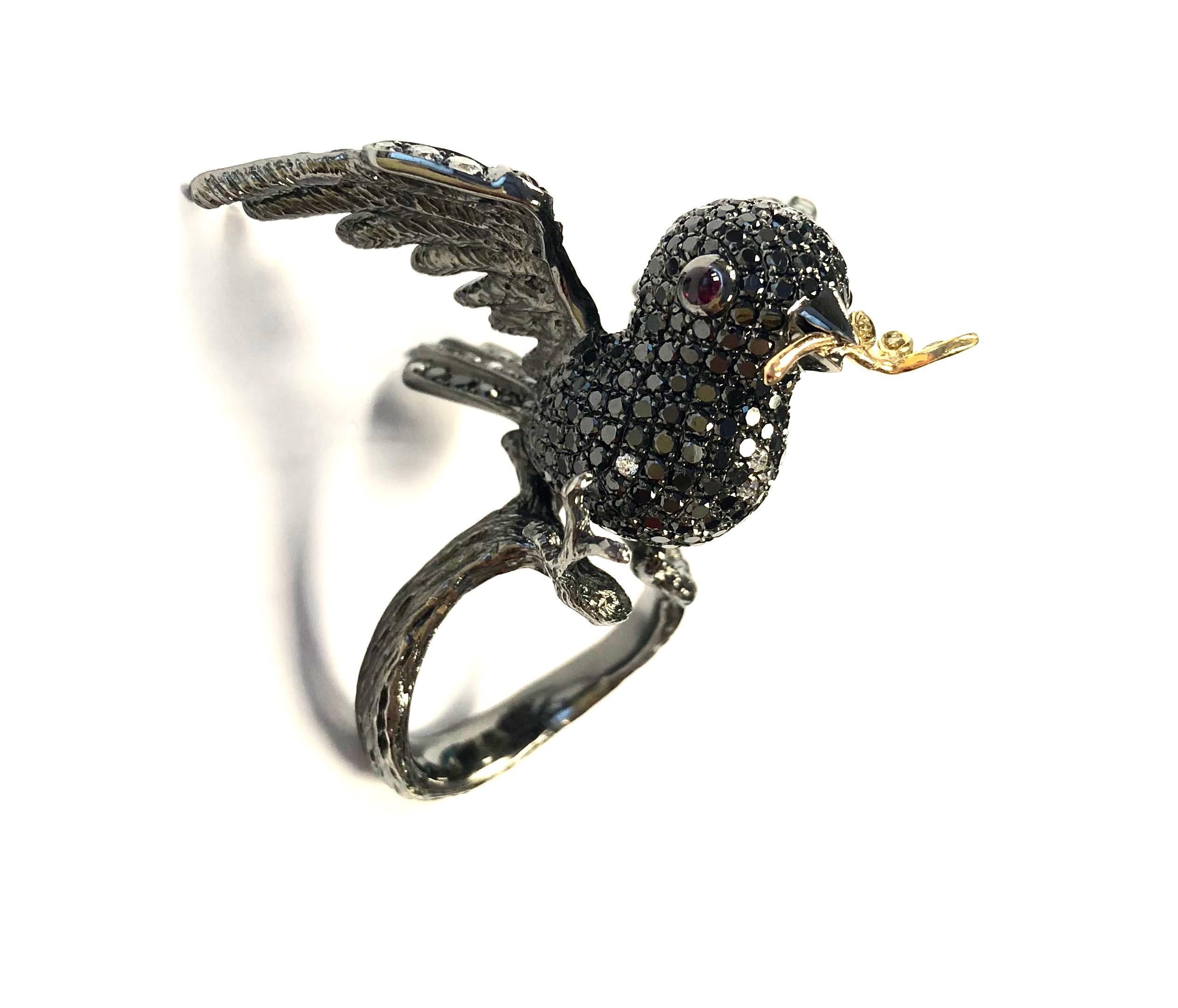 One-of-a-kind 18K gold ring, featuring a bird in flight holding a yellow diamond accented branch in its beak. The bird is set with black and white diamonds, accented by ruby cabochon eyes. The bird and the band are made of blackened gold, the branch