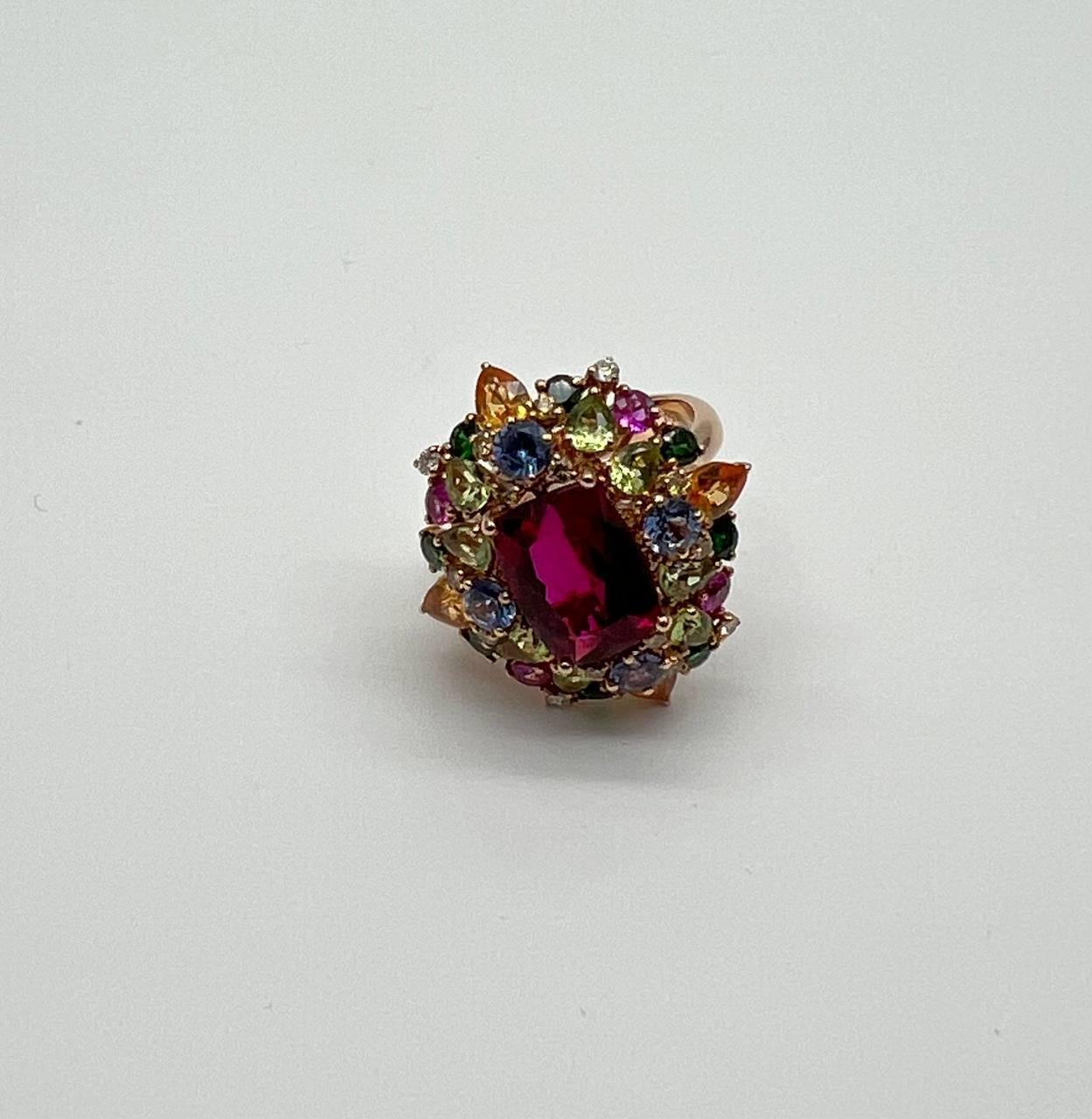 Questo squisito pezzo presenta una vibrante serie di gemme multicolori incastonate in una lussuosa fascia d'oro. Al centro, un grande e affascinante rubino rosso è circondato da un'aureola di pietre più piccole nei toni del blu, verde, giallo e