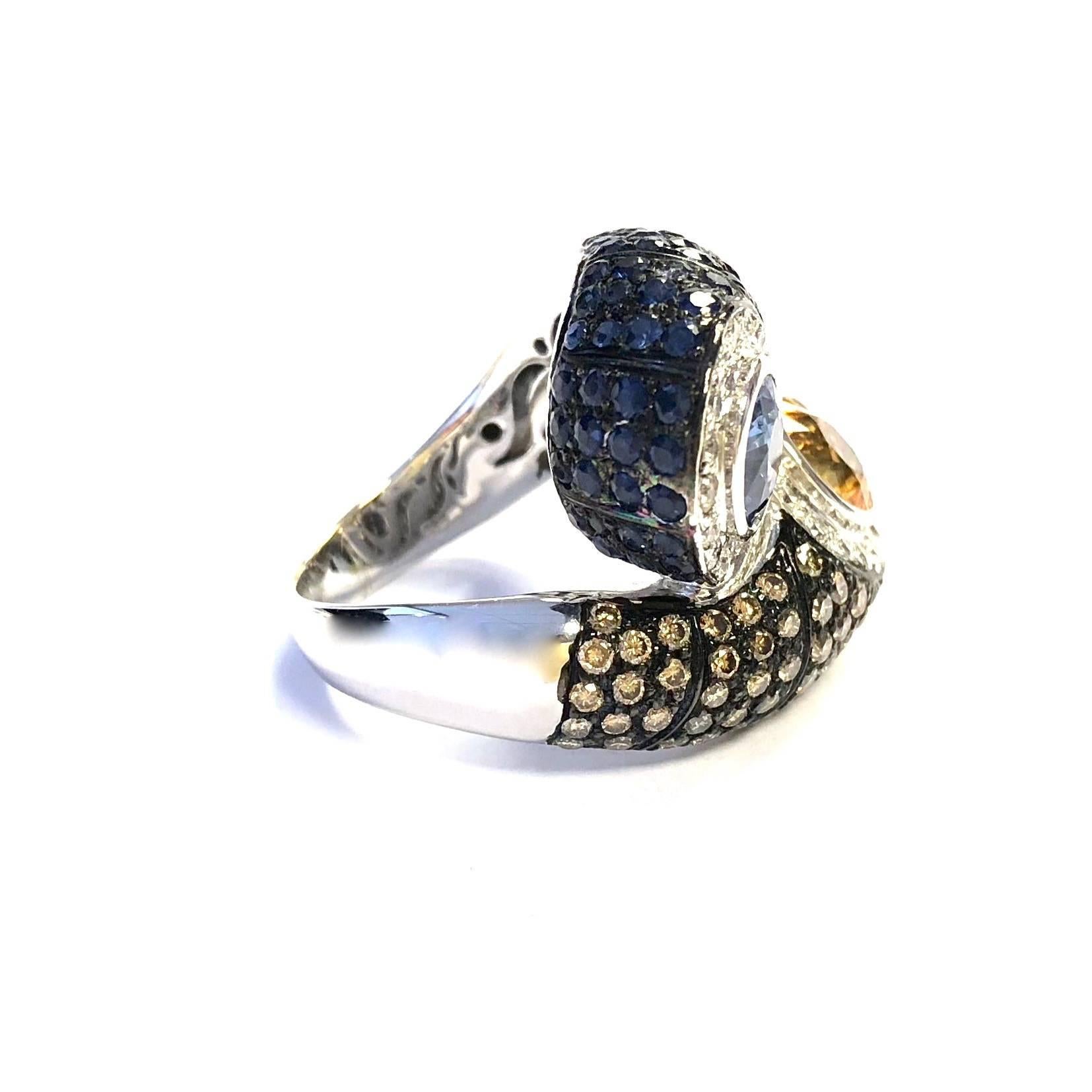 Crafted in 18K white gold, the ring fetures an oval yellow sapphire set within a bezel of white diamonds, supported by a gallery of brown diamonds, and a blue sapphire set in white diamonds, supported by a gallery of blue sapphires. Fully finished