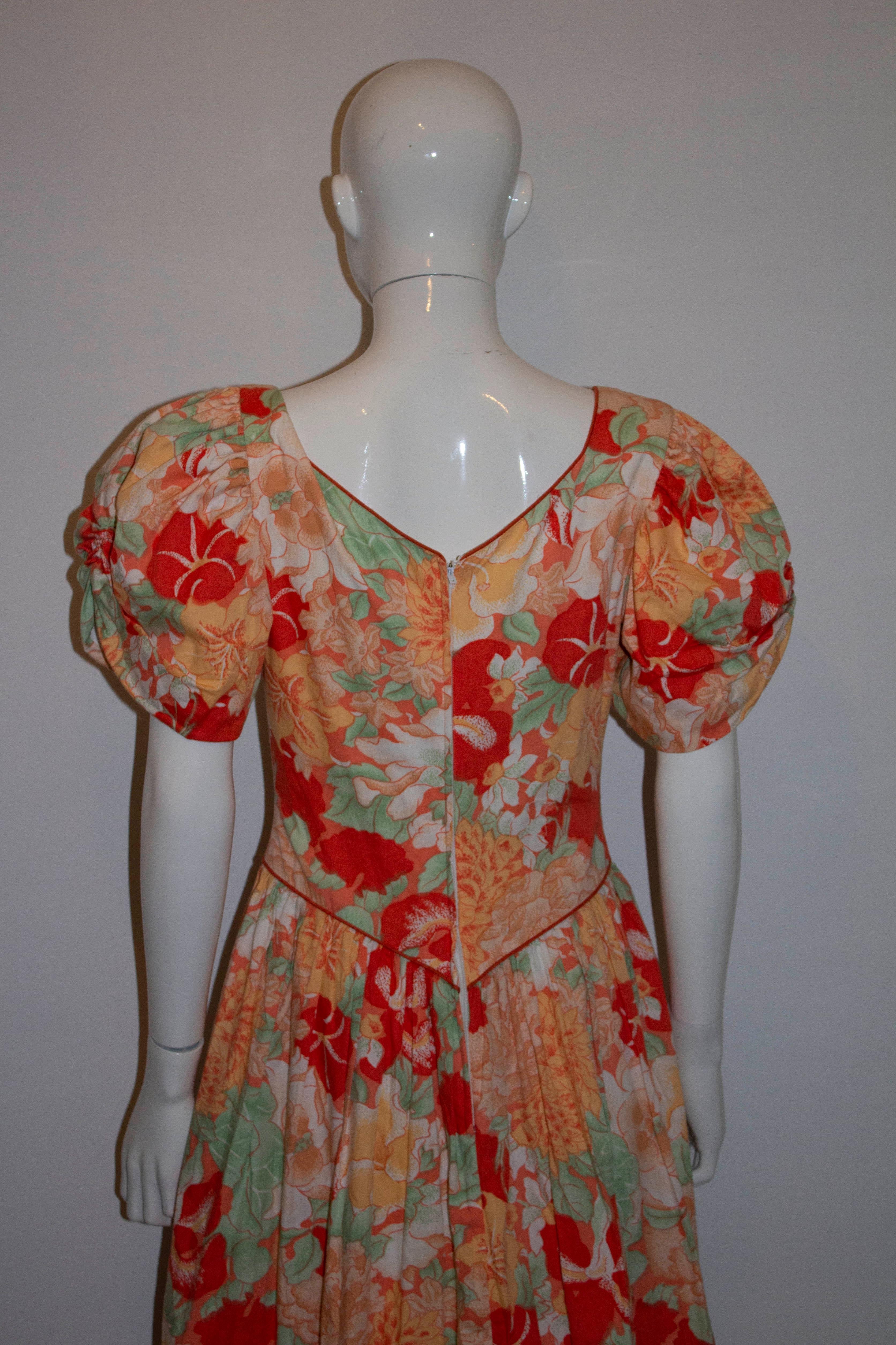 Une magnifique robe florale vintage en coton dans un mélange d'orange, de rouge et de vert. La robe a des manches bouffantes et un décolleté en forme de cœur avec une fermeture éclair centrale au dos.  Il est doublé en coton et possède une jupe